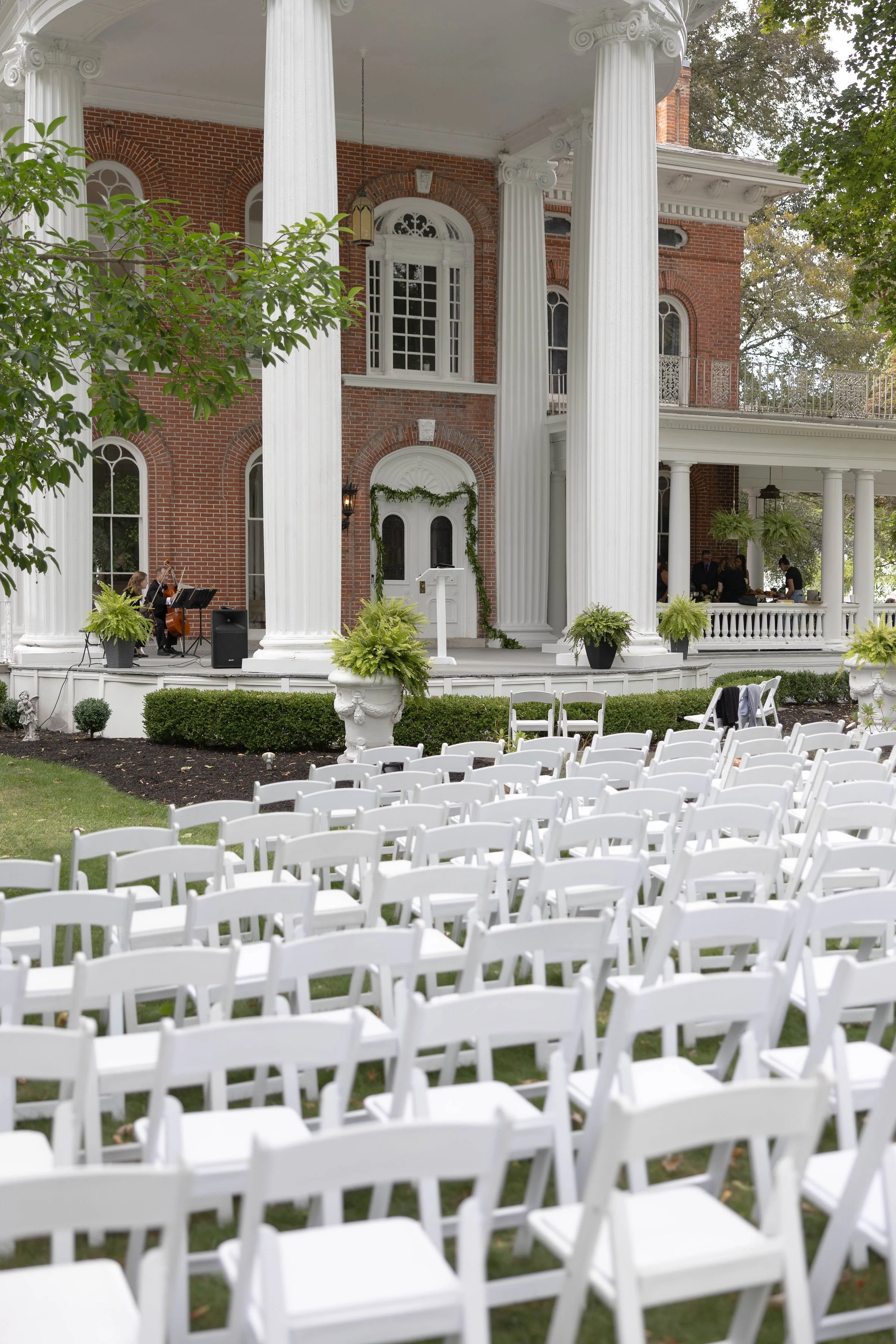 An outdoor wedding setup with a large red brick mansion, white columns, and white chairs arranged on the grass. Musicians are playing on the porch, and guests are seated, preparing for a ceremony.