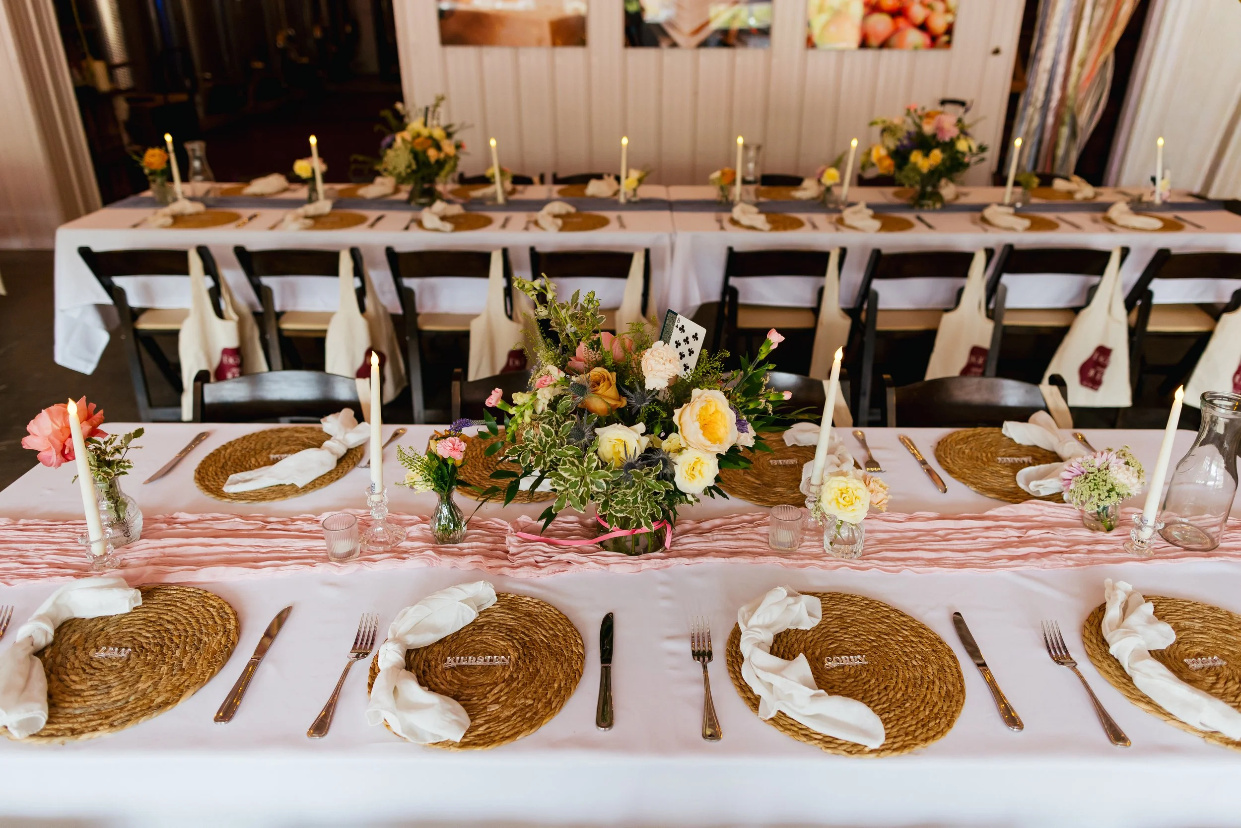 Wedding reception table decorated with a pink tablecloth, floral centerpieces, candles, and place settings with woven placemats and cloth napkins.