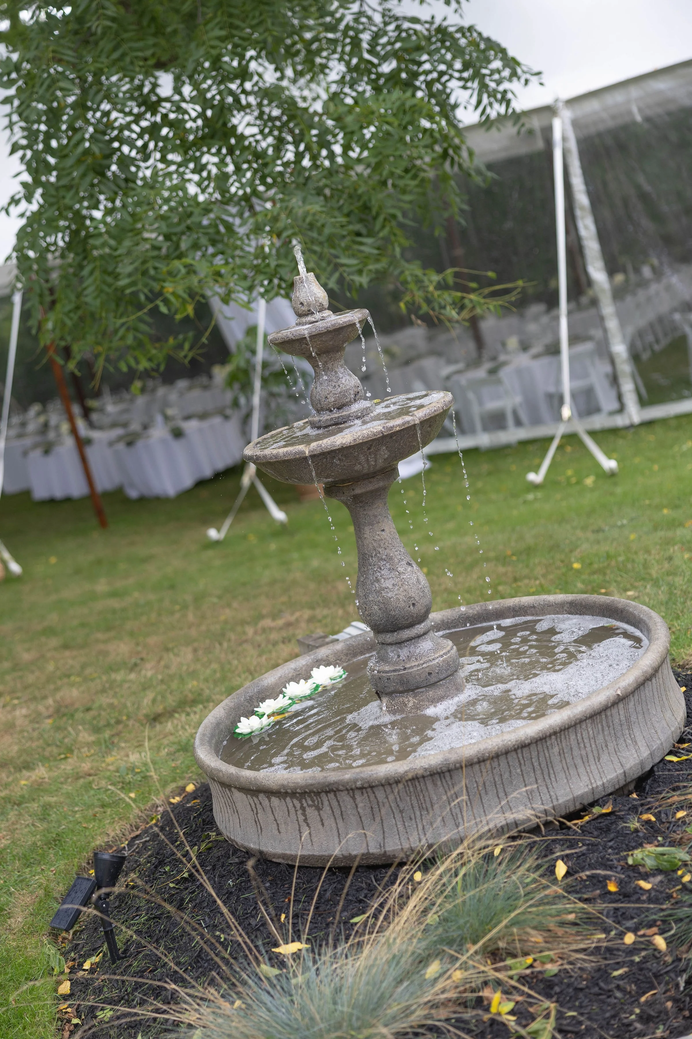 A three-tier stone fountain with water flowing from the top tier to the lower basins, situated in a landscaped flower garden with grass, ivy, and plants. In the background, there are trees and a white fence.
