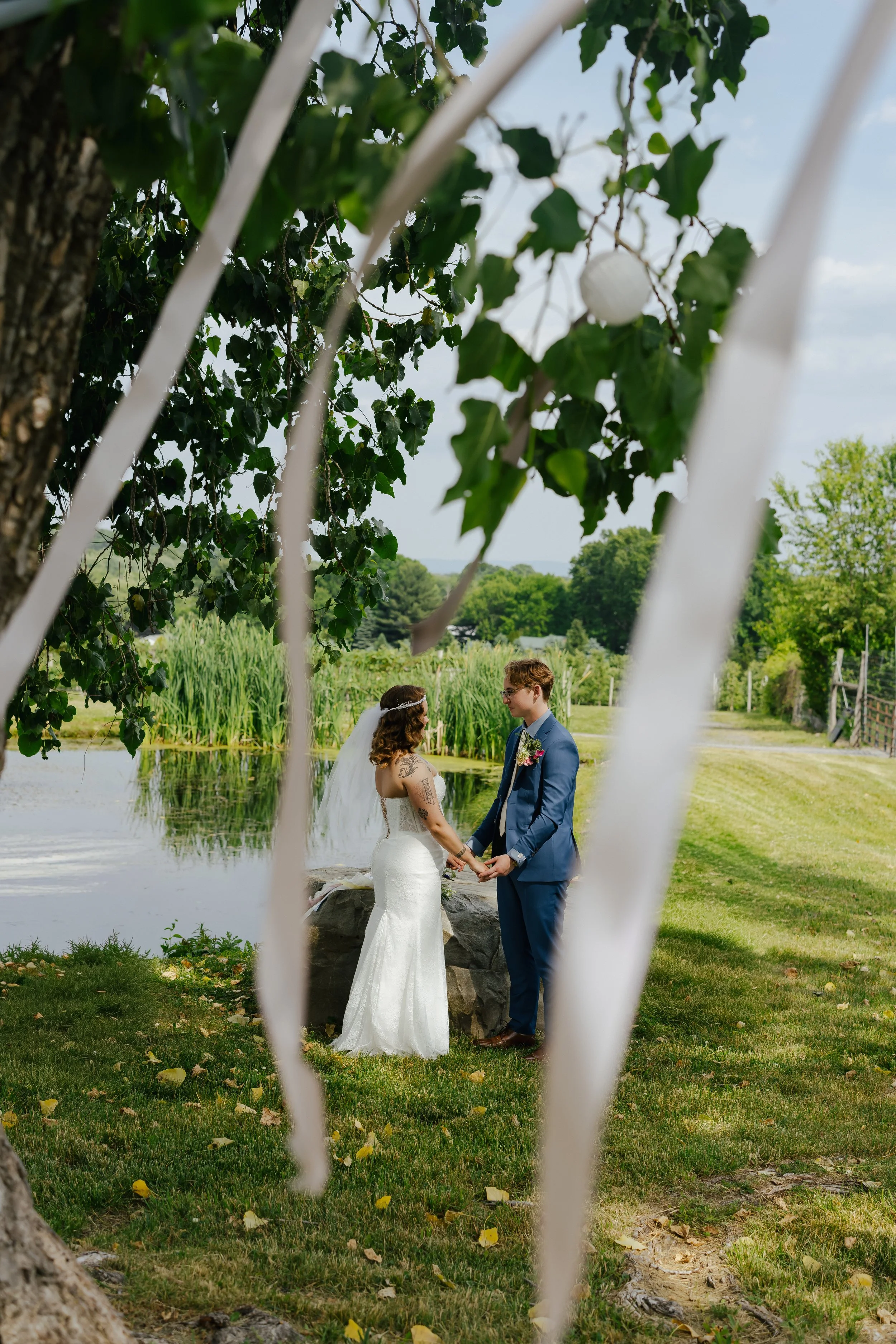 A bride and groom holding hands during an outdoor wedding ceremony near a pond, seen through white lattice. Trees with green leaves and a blue sky are in the background.