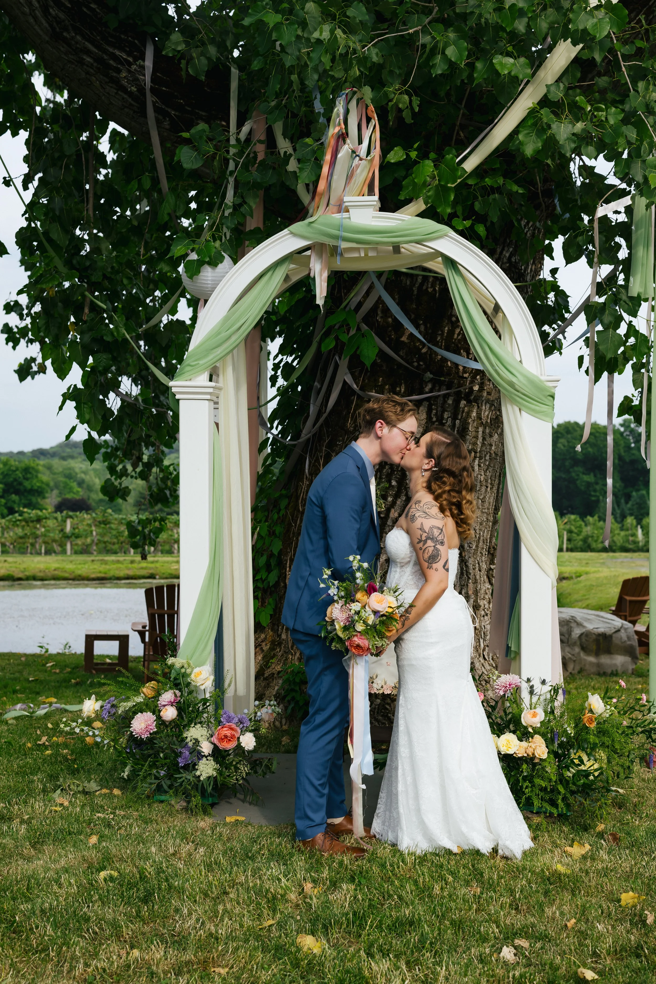 A bride and groom share a kiss during their outdoor wedding ceremony under a decorative arch adorned with draped fabric and flowers. They are standing in front of a large tree with greenery and open fields in the background.