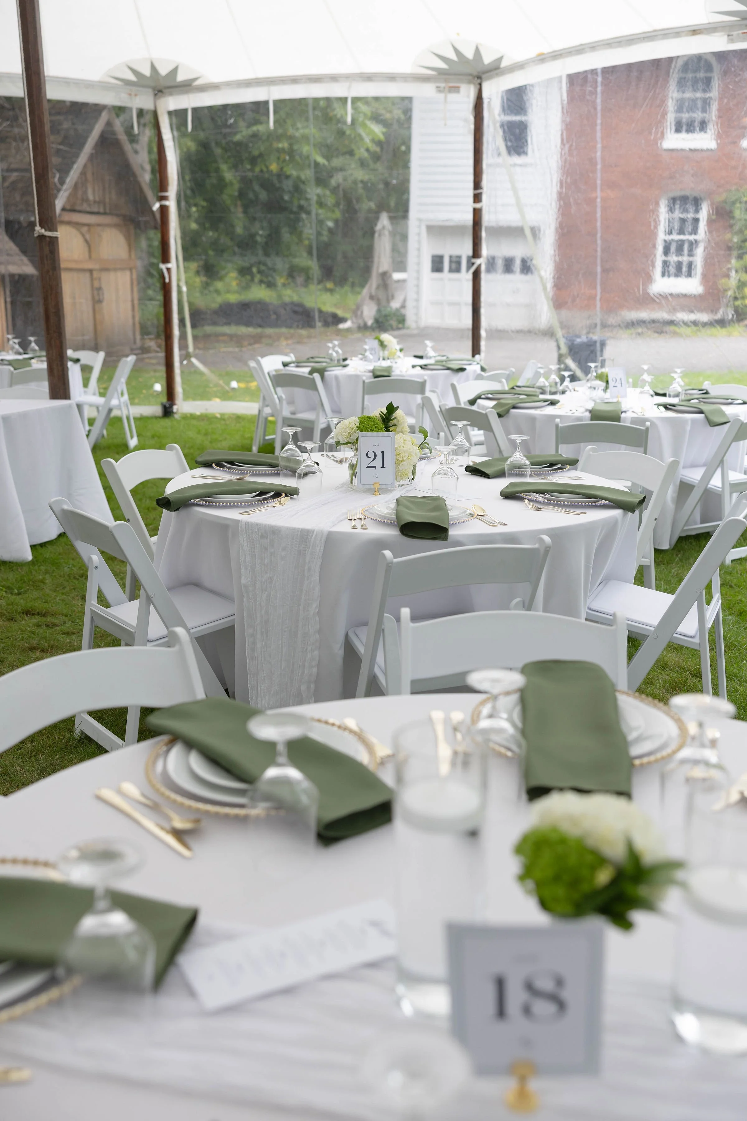 Round tables set for a formal event under a white tent outdoors, with white chairs, green napkins, floral centerpieces, table numbers, and place settings with plates, glasses, and silverware.
