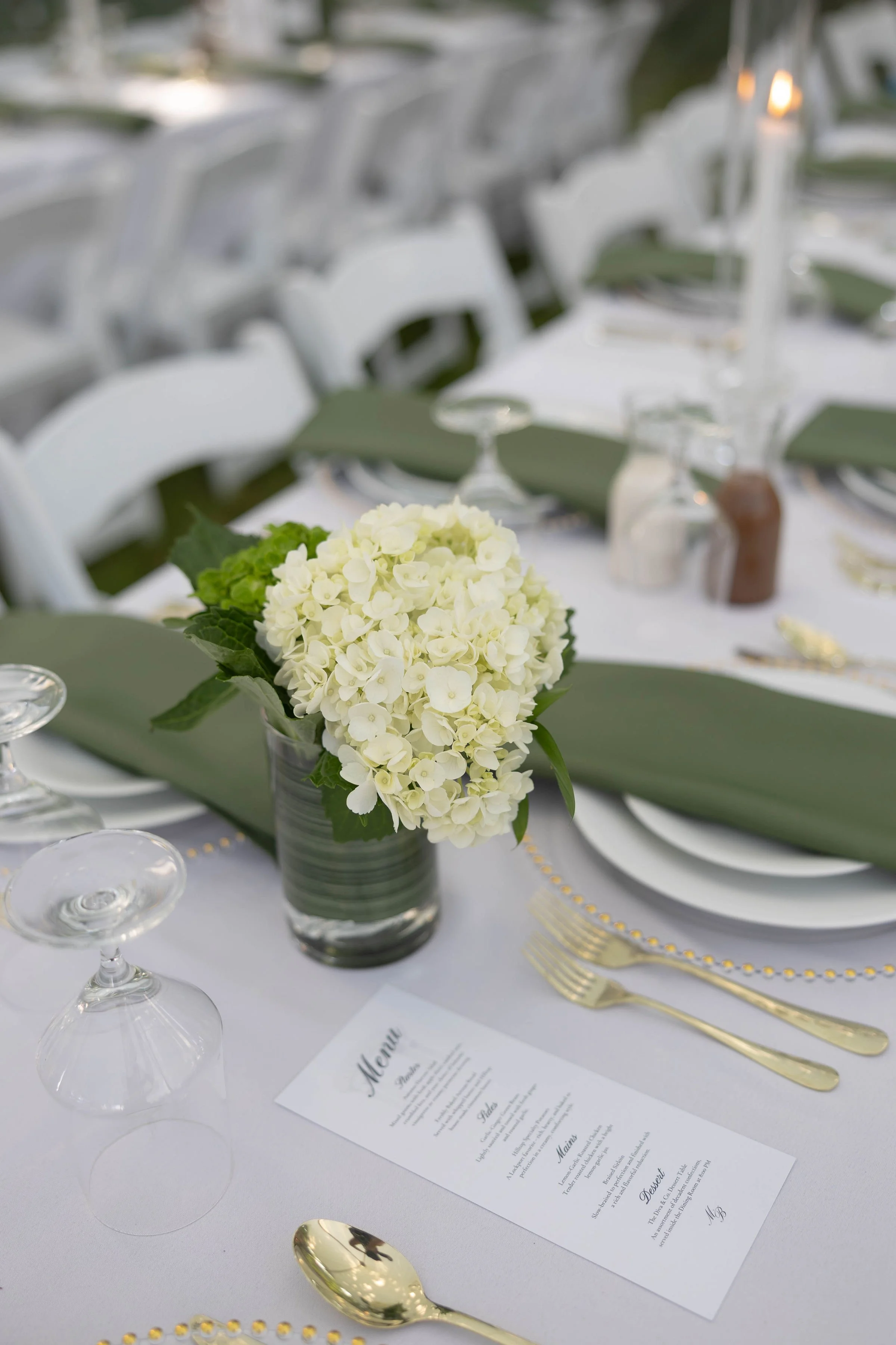 Elegant table setting at a wedding or formal event, with a white tablecloth, gold cutlery, a floral centerpiece of white hydrangeas, wine glasses, and a printed menu.