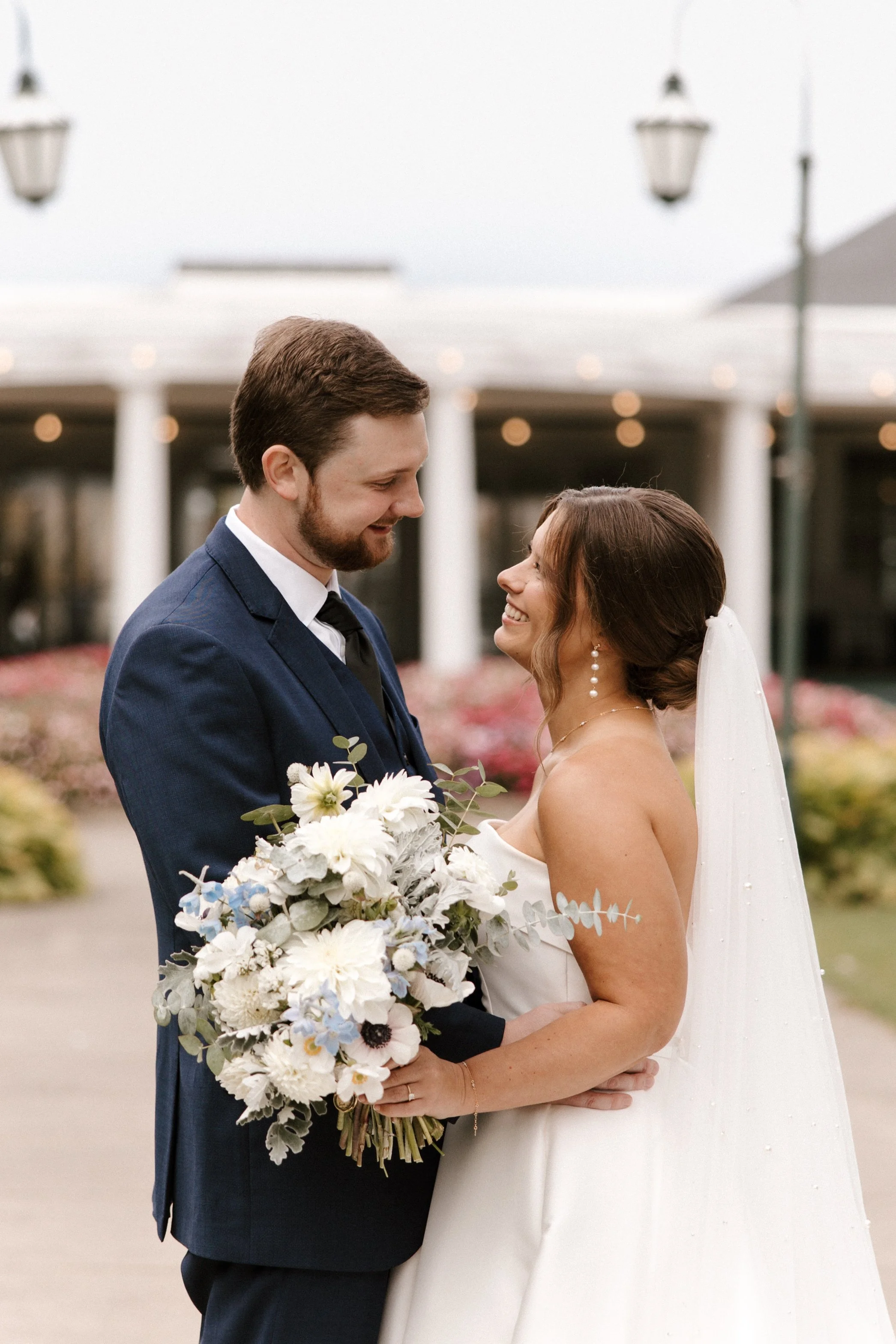 A bride and groom look at each other and smile during their wedding ceremony or photoshoot outdoors, with a background of white columns, hanging lamps, and a cloudy sky. The groom wears a blue suit and the bride wears a white wedding dress and veil, 