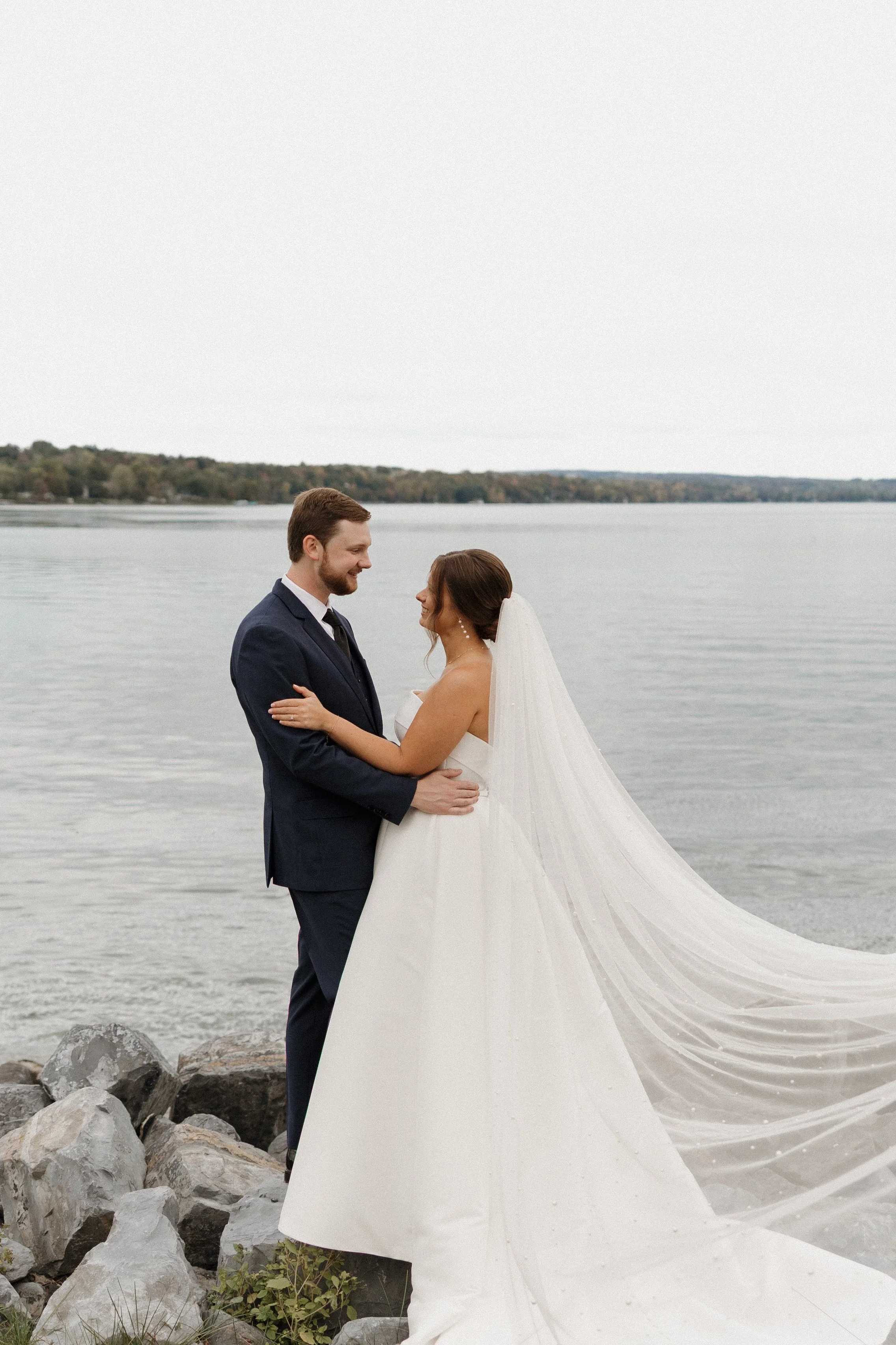 A bride and groom holding each other and smiling on a rocky shoreline by a lake, with the bride wearing a white wedding dress and veil, and the groom wearing a dark suit, against a backdrop of water and distant trees under an overcast sky.