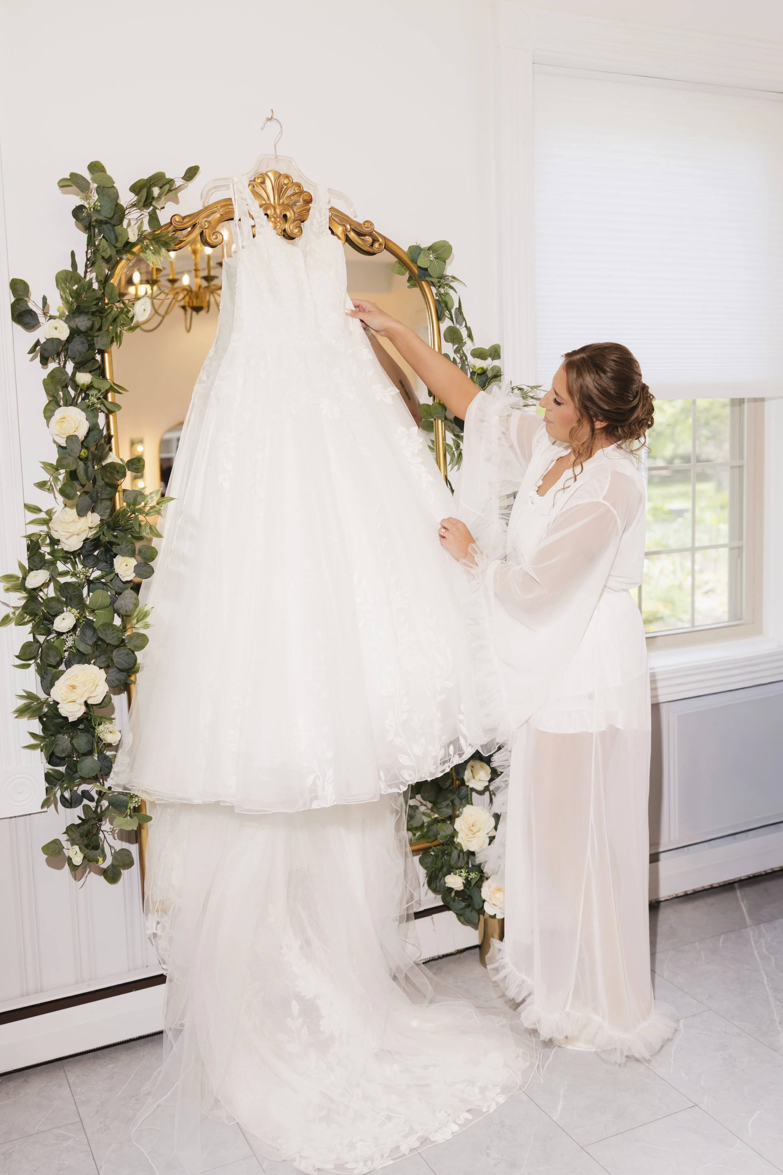 A woman dressed in a white robe is holding a wedding dress on a hanger in front of a mirror decorated with green leaves and white roses. The wedding dress is hanging on the mirror, which has a gold frame, and there is a chandelier reflected in the mi