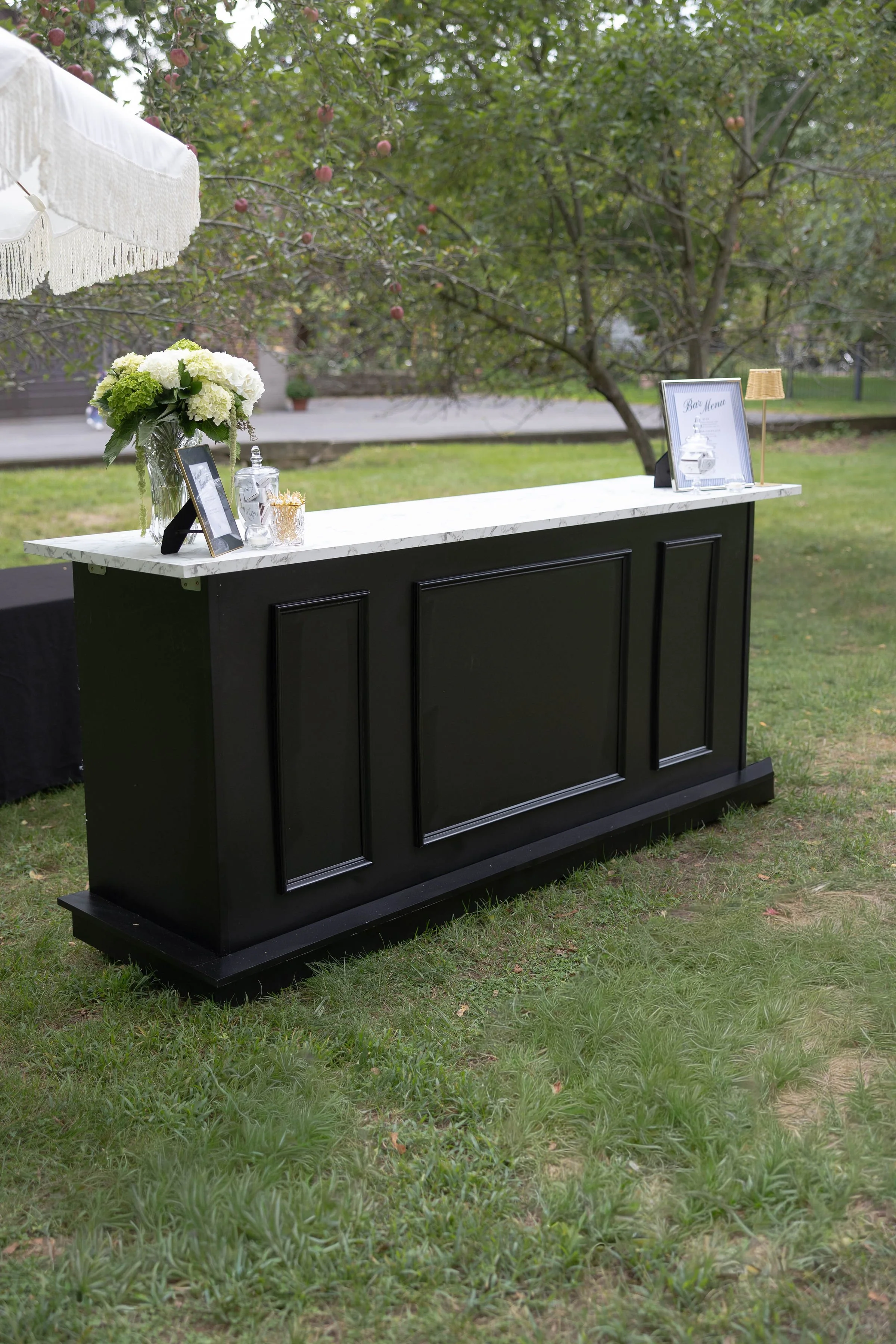 Outdoor bar setup with a black bar counter with a white marble top, decorated with a vase of white and green flowers, framed menu, glass containers, and a small lamp, set in a grassy area with trees and apple branches in the background.