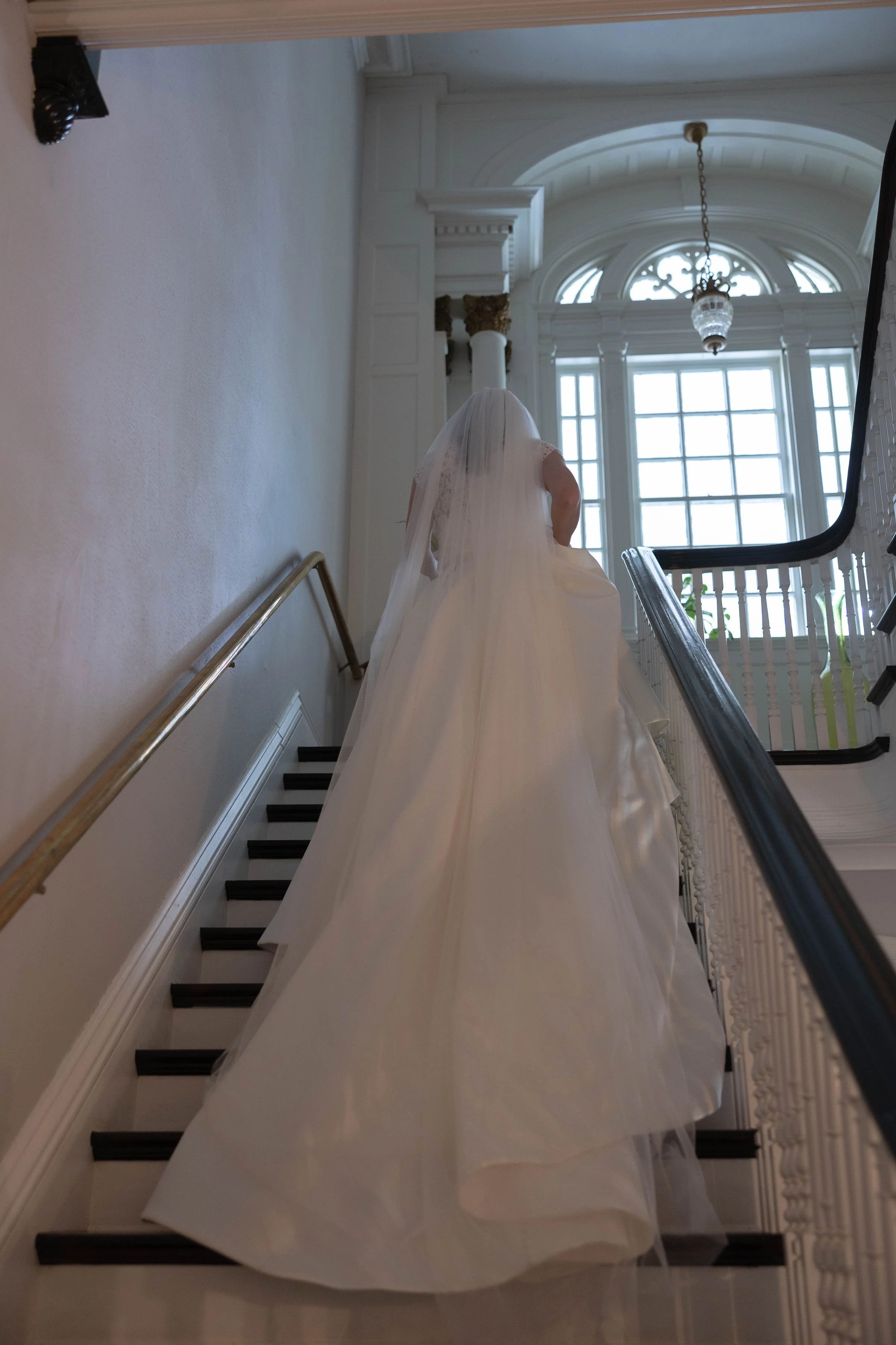 A bride in a white wedding dress with a long train ascending a stairway inside a grand building with high ceilings, large arched windows, and elegant architectural details.