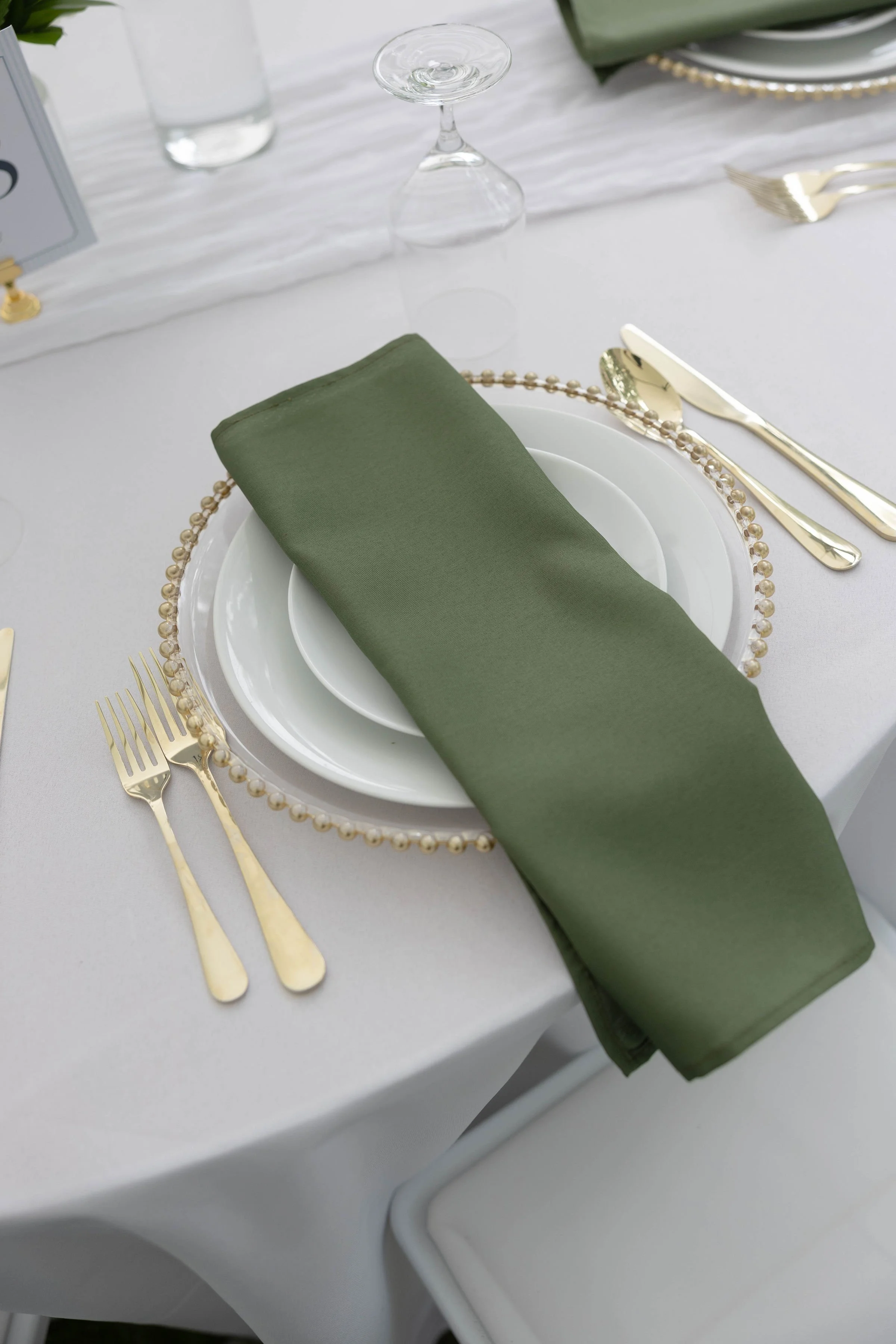A formal table setting with a white tablecloth, a white plate layered with a smaller white bowl, a green cloth napkin, gold flatware, a pearl-edged glass plate, and an upside-down clear glass. There is also a water glass and a partial view of a sign 