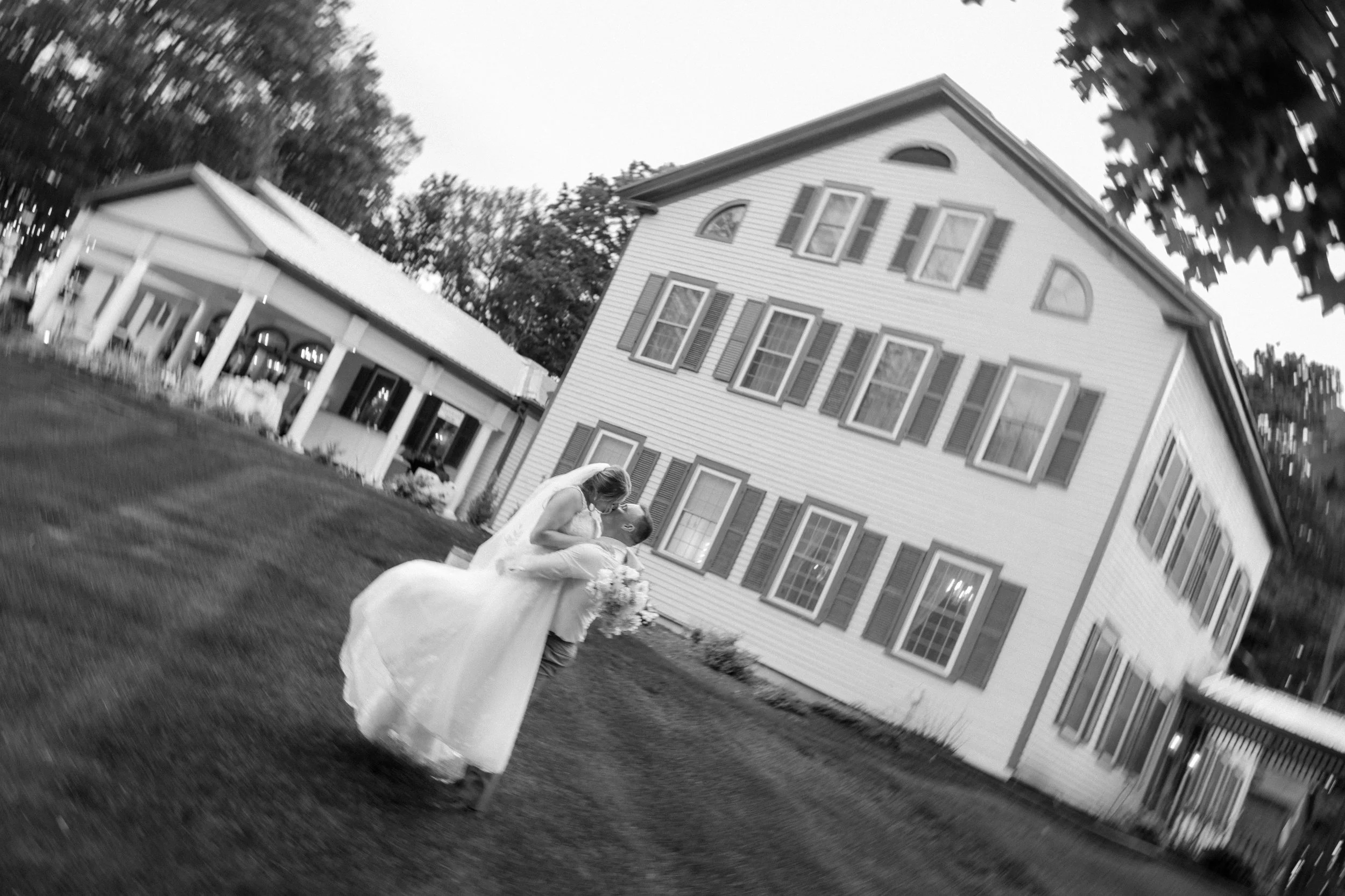 A woman in a wedding dress and a man in a tuxedo sharing a kiss outdoors in front of a large white house with multiple windows and trees, during a wedding celebration.