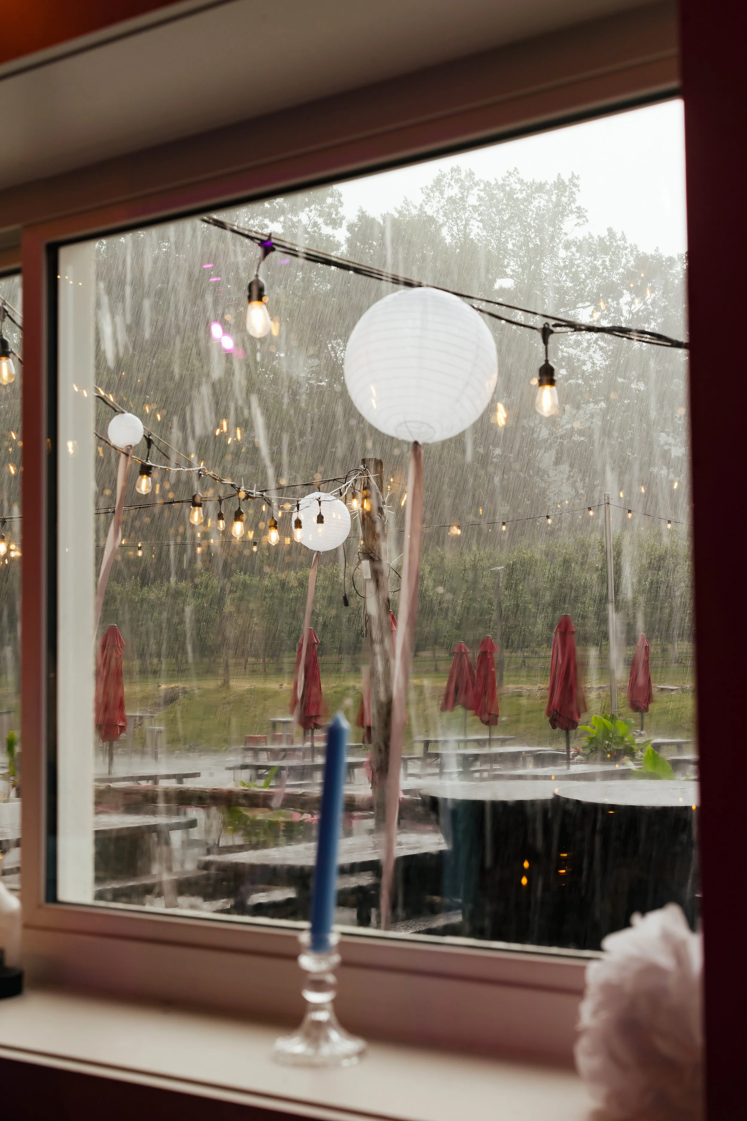 Rain pouring outside through a window decorated with hanging paper lanterns and string lights; red umbrellas lined up outside on a patio or garden area.
