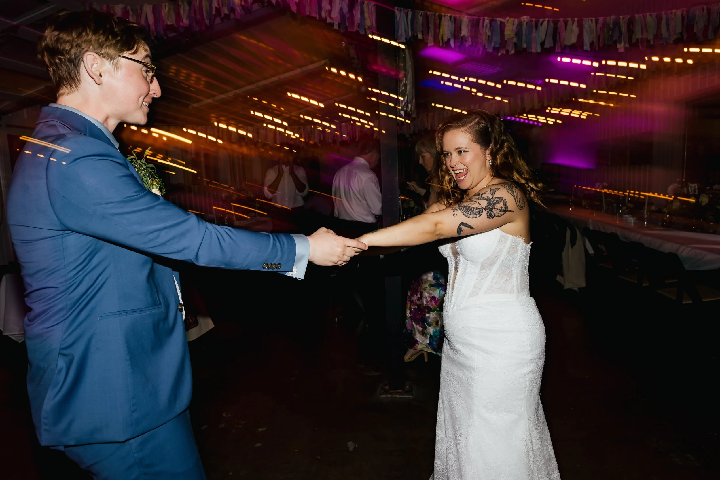 Couple dancing at a wedding reception, with the man in a blue suit and the woman in a white wedding dress, smiling and holding hands.