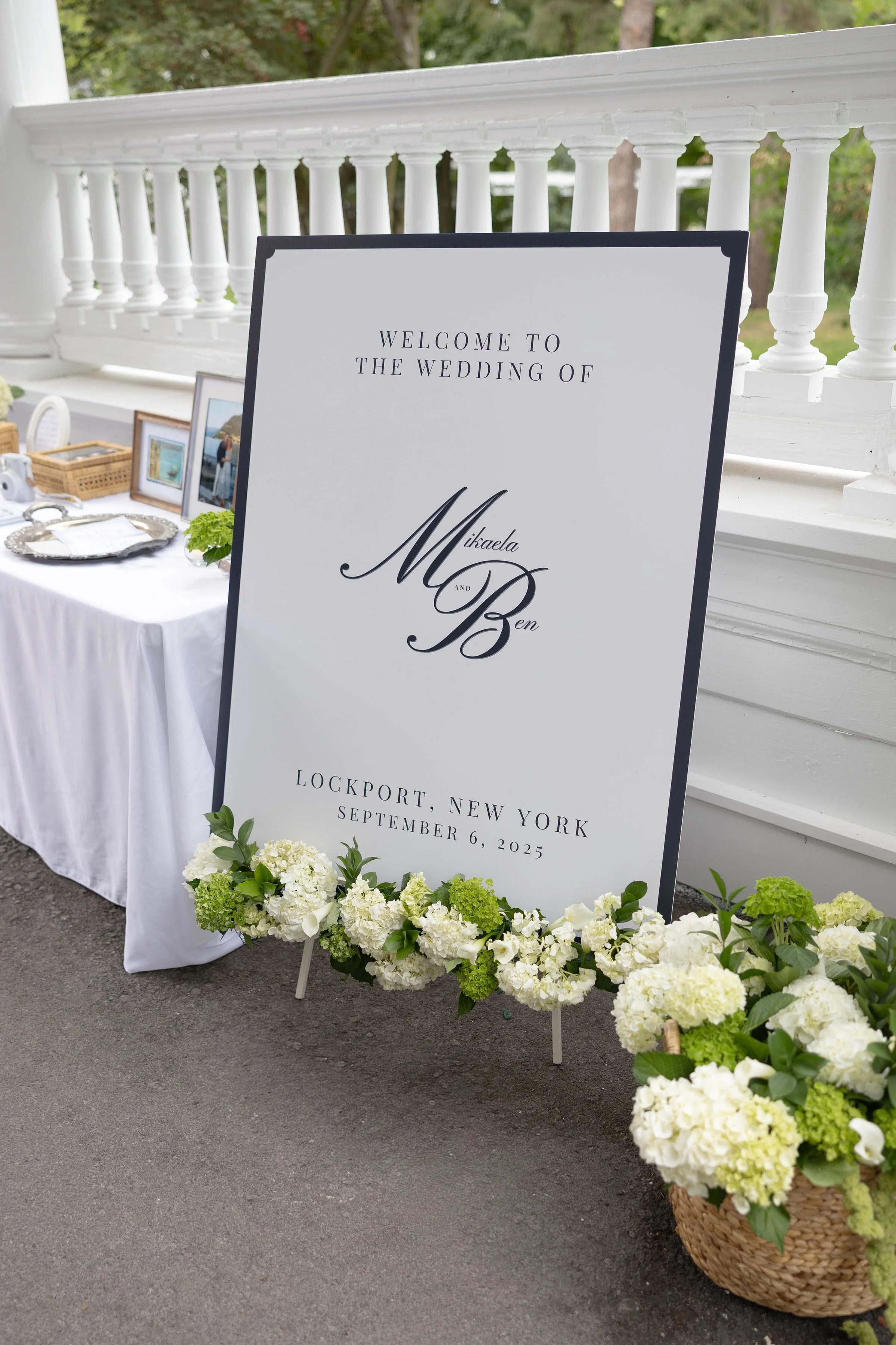 Wedding welcome sign on a white easel with floral decorations, reading "Welcome to the wedding of Mikaela and Ben, Lockport, New York, September 6, 2025," surrounded by white and green hydrangeas and other flowers in baskets.