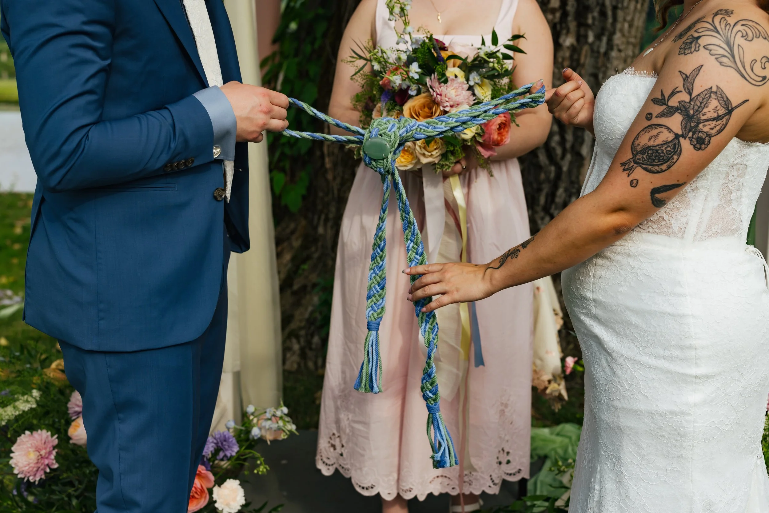 A wedding ceremony where the bride and groom are holding a braided cord as part of a unity ritual, with a bridesmaid holding a bouquet in the background. The bride has tattoos on her arm, wearing a white lace dress, and the groom is in a blue suit.