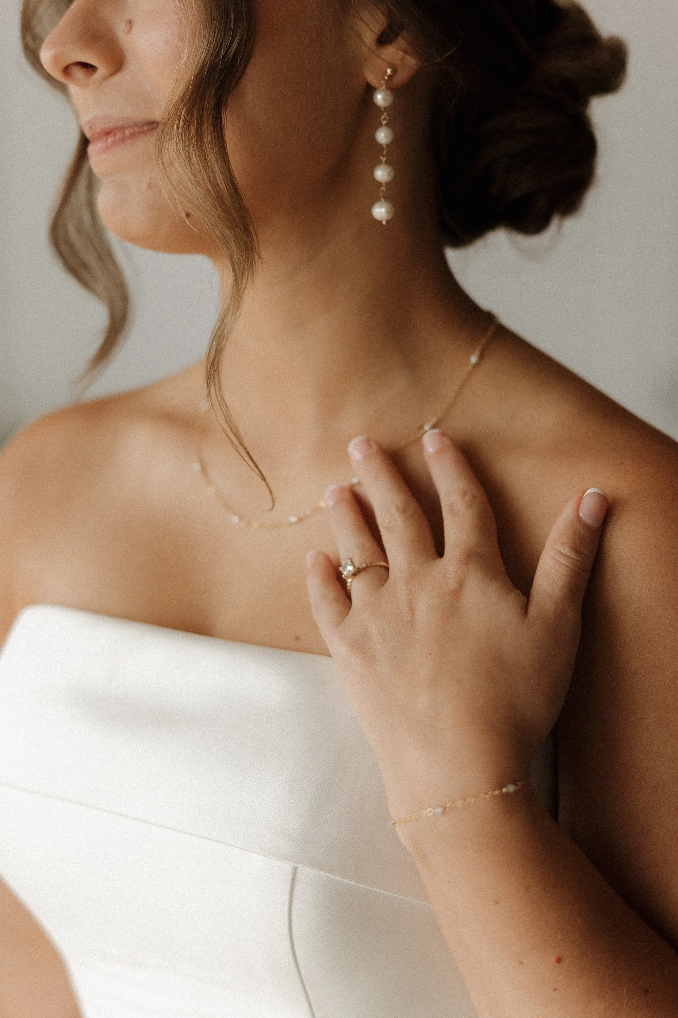 Close-up of a woman wearing pearl earrings, a delicate necklace, and a bracelet, with her hand resting on her chest, facing left, against a plain background.