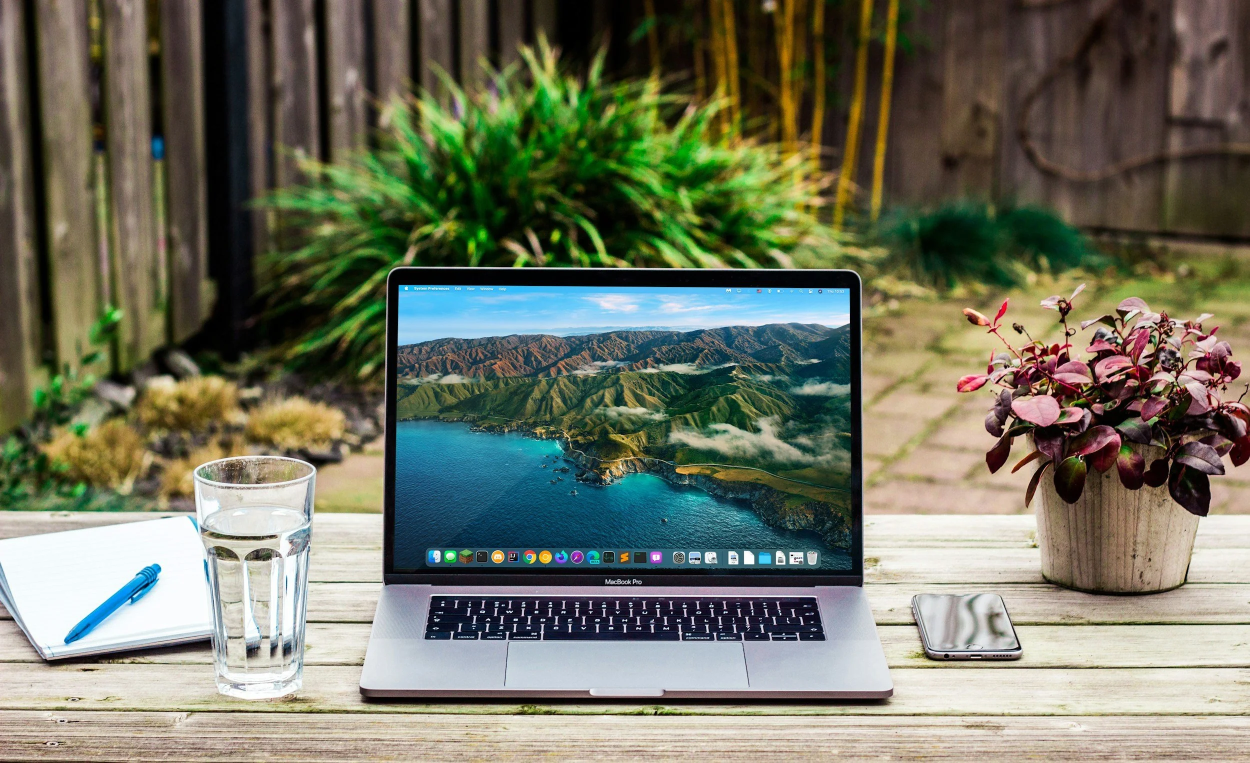 A MacBook Pro laptop on a wooden outdoor table displaying a mountain landscape on its screen, with a glass of water, a notebook with a blue pen, and a smartphone nearby. In the background, there are green and purple plants and a wooden fence.