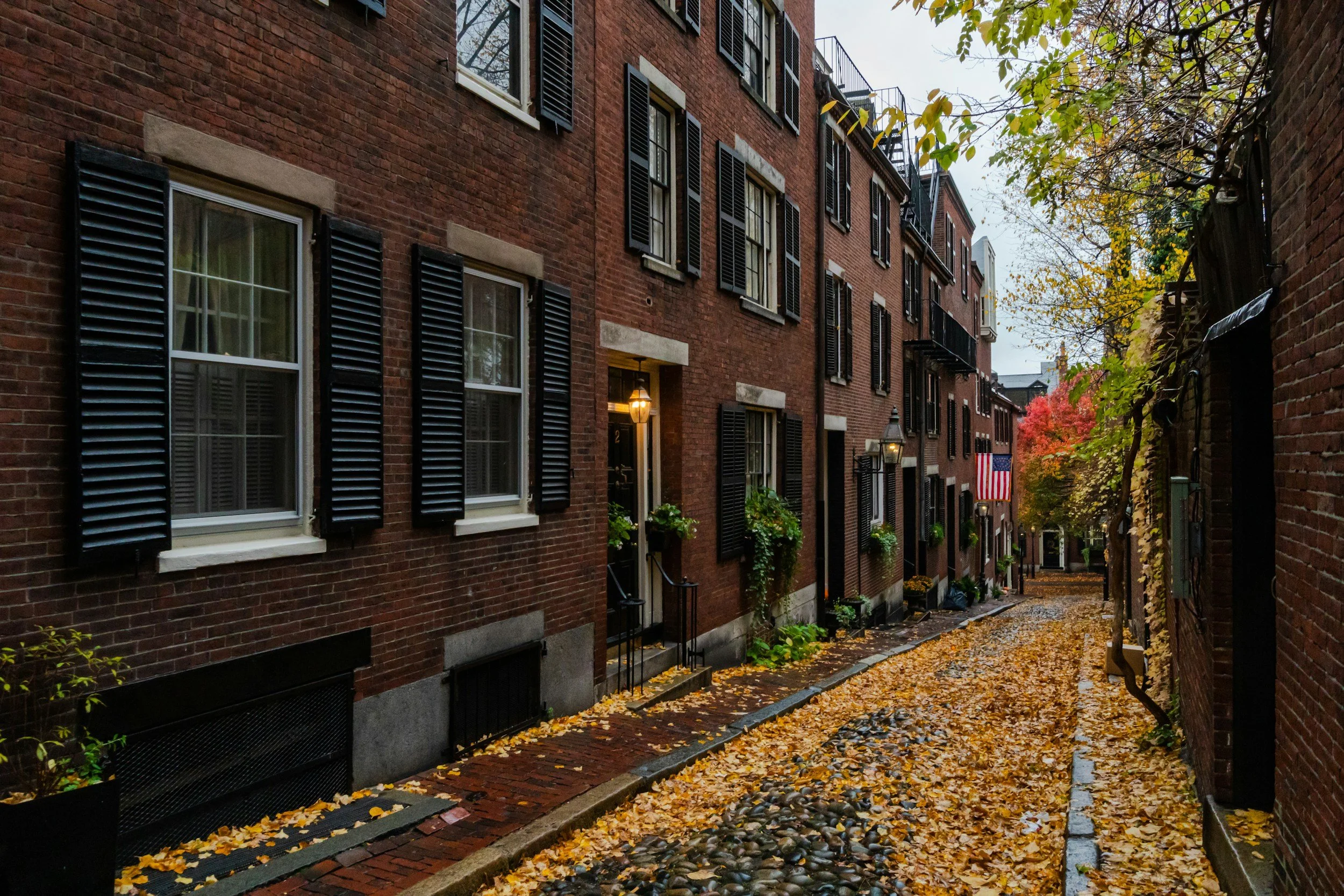 A narrow brick alleyway with fallen yellow leaves on the brick and cobblestone ground, flanked by red brick apartment buildings with black shutters and ivy, and decorated with street lamps and an American flag, during autumn.