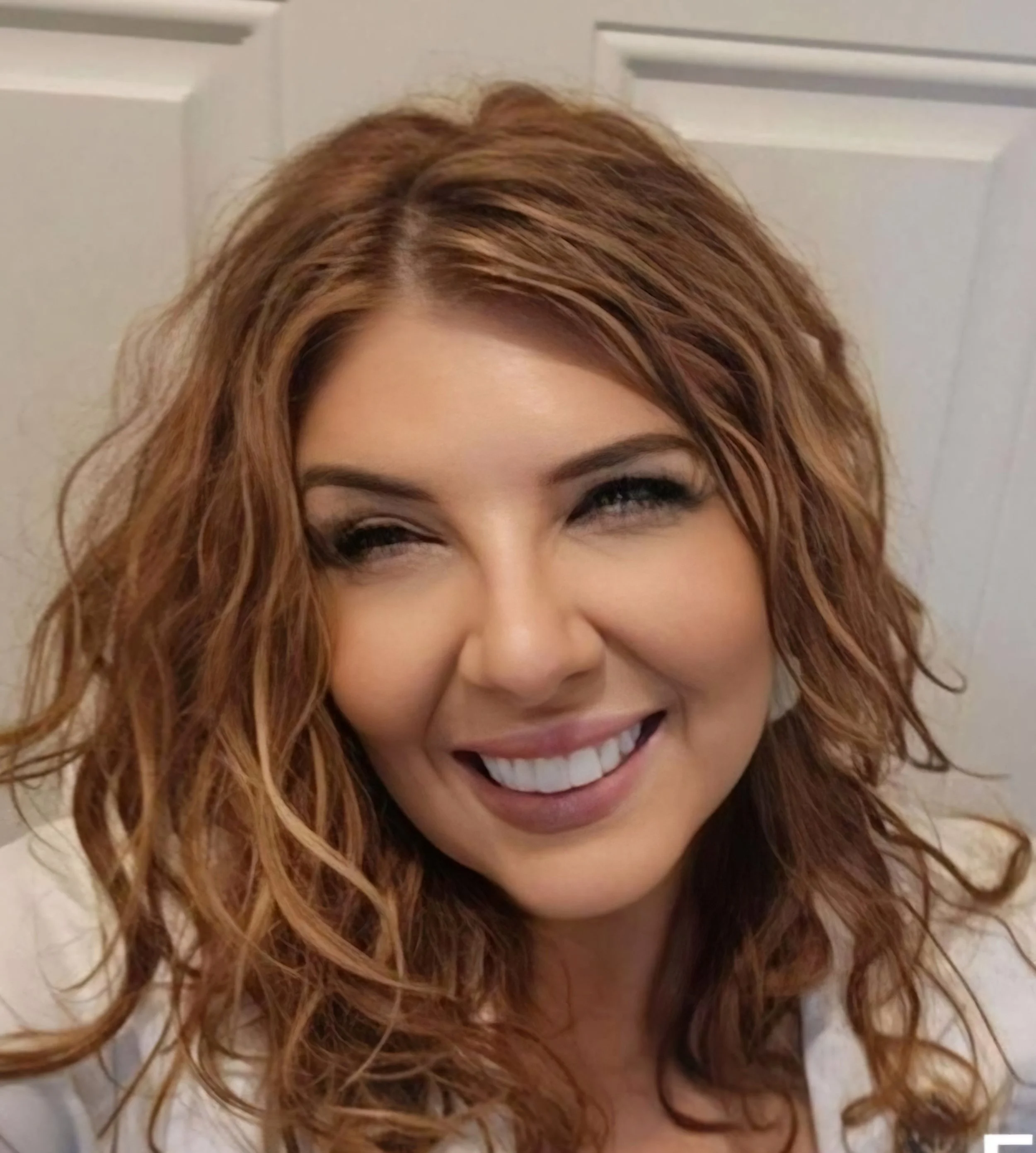 Close-up of a smiling woman with wavy red hair, light skin, and makeup, posing indoors against a white wall.