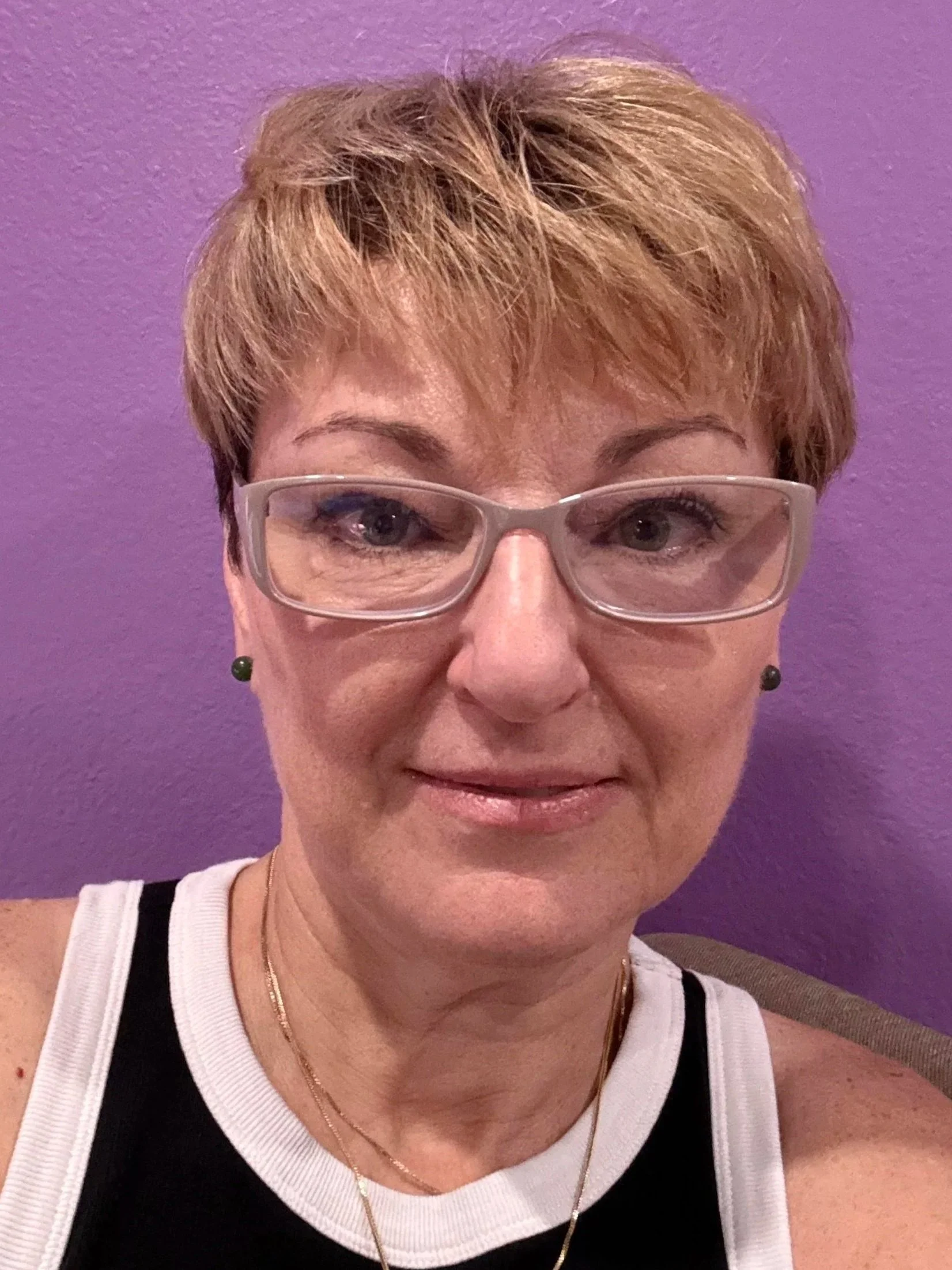 A woman with short brown hair, wearing clear glasses, a black and white top, and gold necklaces, standing against a purple wall.