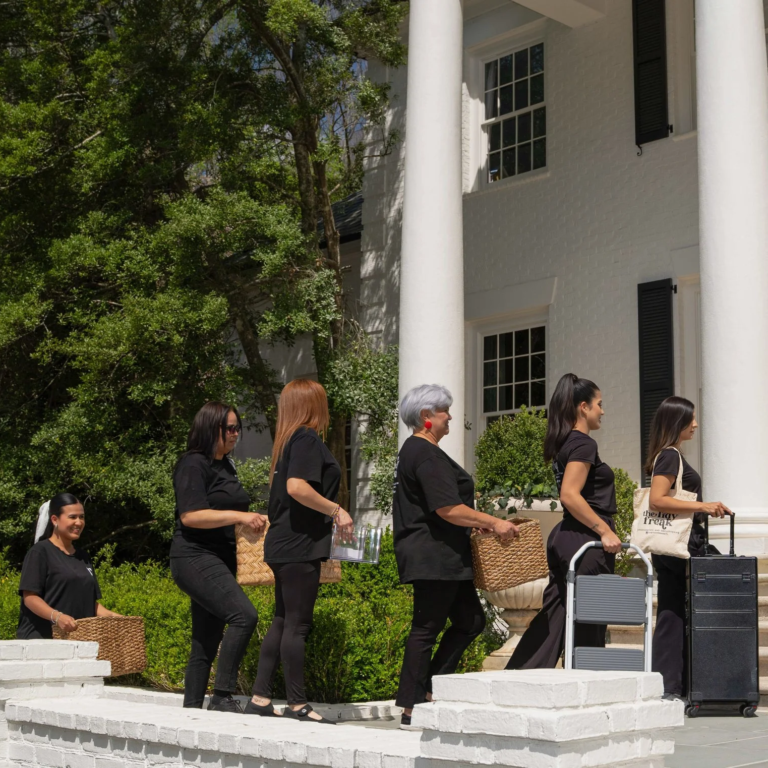 A group of women walking in a line with some carrying baskets and luggage outside a white house with black shutters, green bushes, and trees in the background.