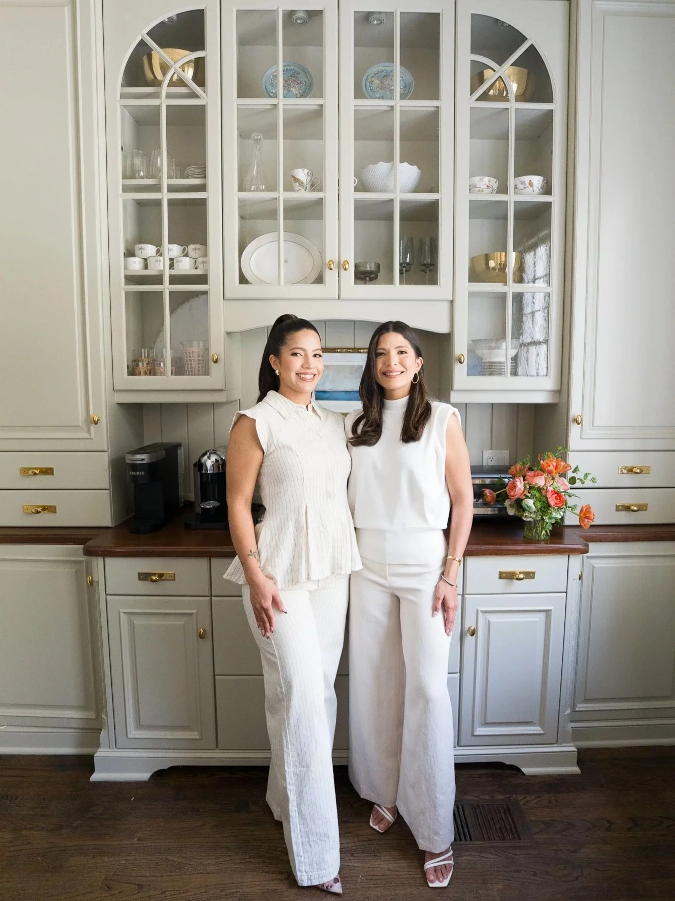 Two women standing in front of a cream-colored kitchen cabinet with glass doors. They are smiling and dressed in white outfits. A desk with flowers, coffee machine, and glasses are visible below the cabinet.