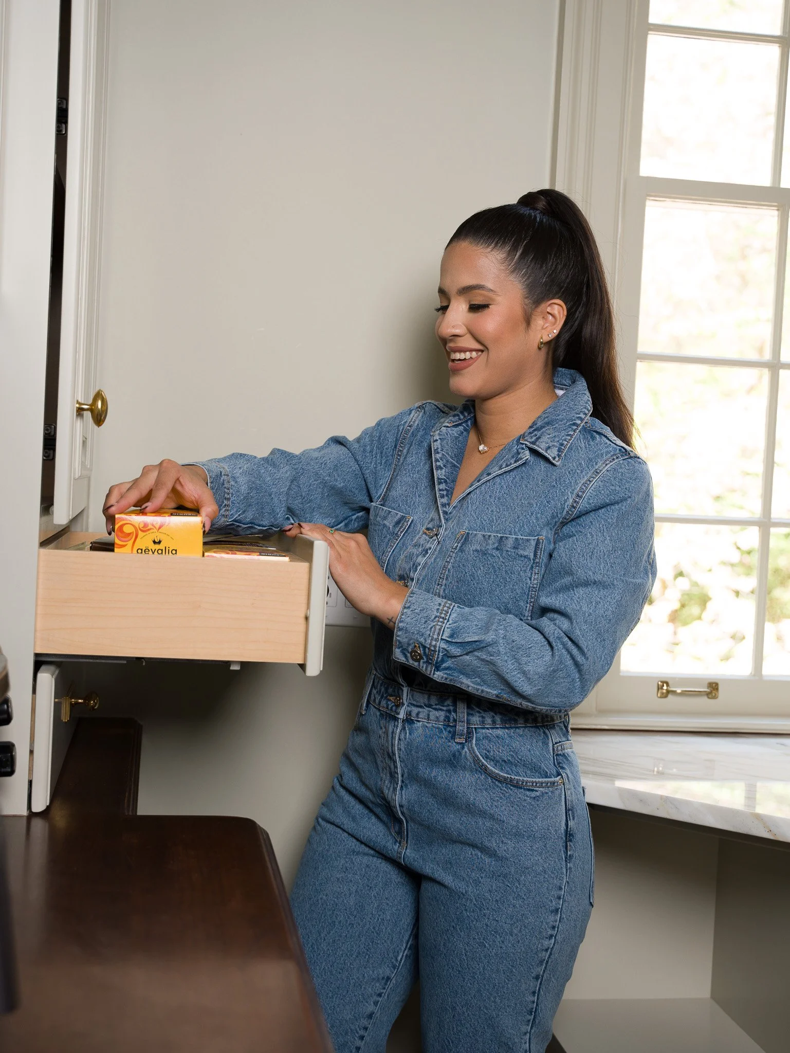A woman with long dark hair in a high ponytail opening a wooden drawer in a white cabinet near a window, wearing a denim jacket and jeans, with a smile on her face.