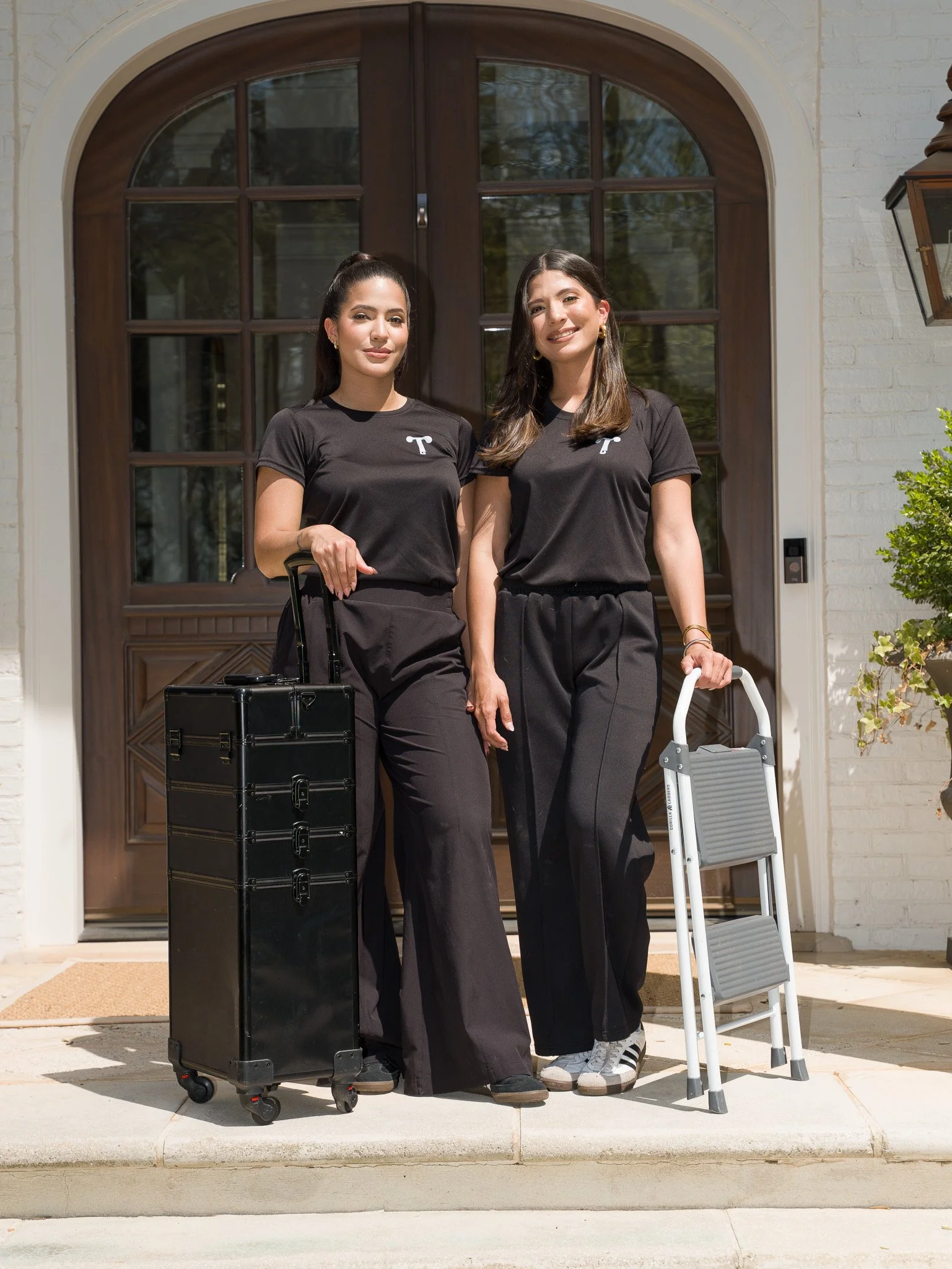Two women standing in front of a house with a wooden door, holding rolling luggage and a step ladder, dressed in matching black t-shirts with a white 'T' logo, outdoors on a sunny day.