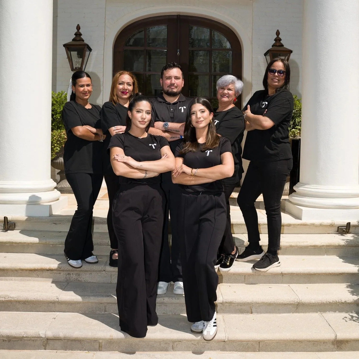 A group of seven people standing on the steps outside a large house with white columns and a wooden door, all wearing black shirts with a white