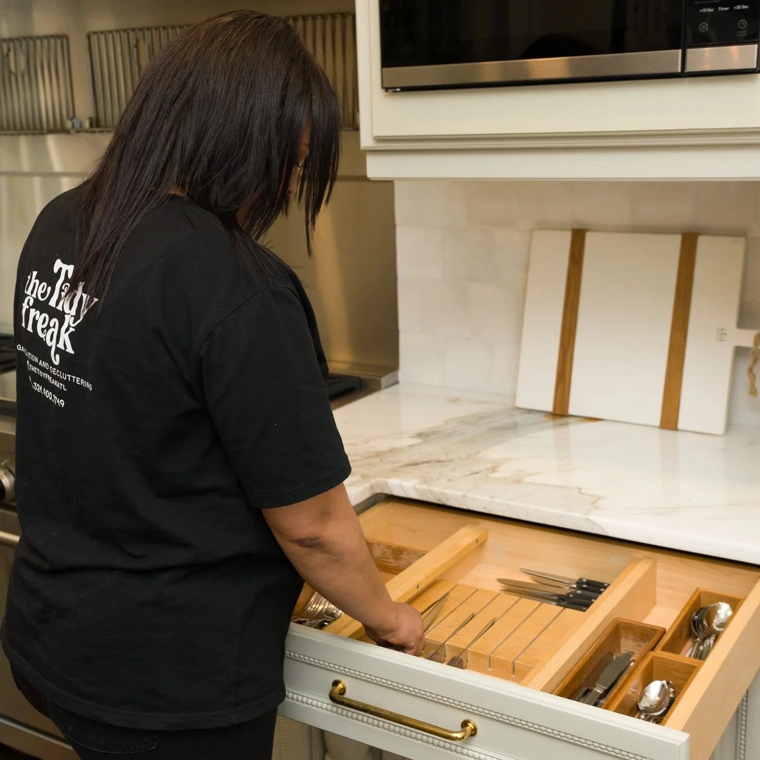 A woman with dark hair wearing a black T-shirt arranging silverware in a wooden tray inside a kitchen drawer.