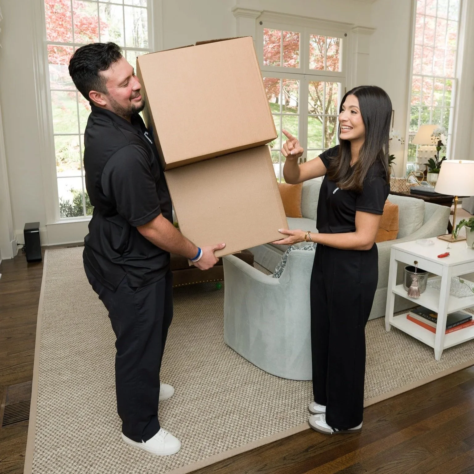 A woman and a man are exchanging large cardboard boxes inside a bright living room with large windows and greenery outside. The woman is smiling and pointing at the man, who is also smiling.