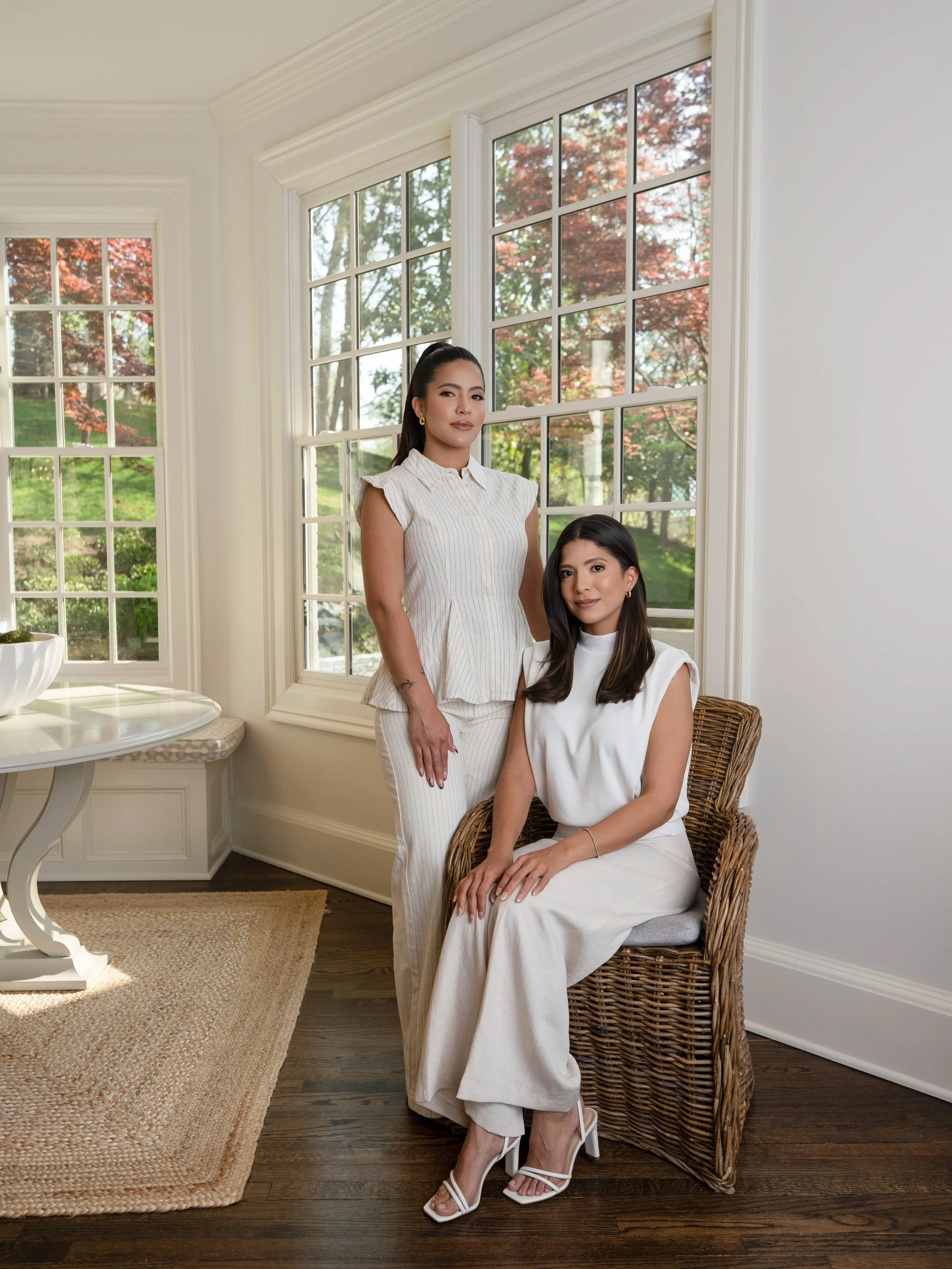 Two women with dark hair and similar facial features in a bright room with large windows and wooden floors. One woman is standing in a sleeveless, white, striped dress with her hair pulled back, and the other is sitting on a wicker chair wearing a sleeveless white outfit with her hair down. Outside the windows, trees with red and green leaves are visible.