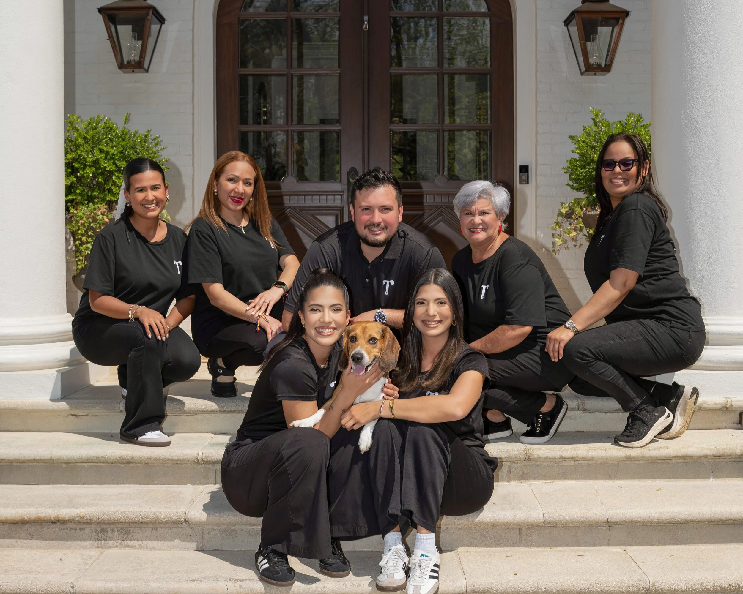 A diverse group of nine people, including eight women and one man, and a dog, posing for a photo on the steps of a house with a white brick exterior, wooden door, and greenery on the sides.