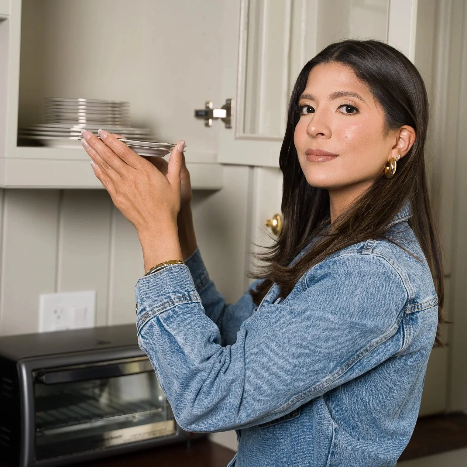Woman with dark hair, wearing a denim jacket and gold earrings, holding a silver plate in a kitchen near a stove and cabinet.