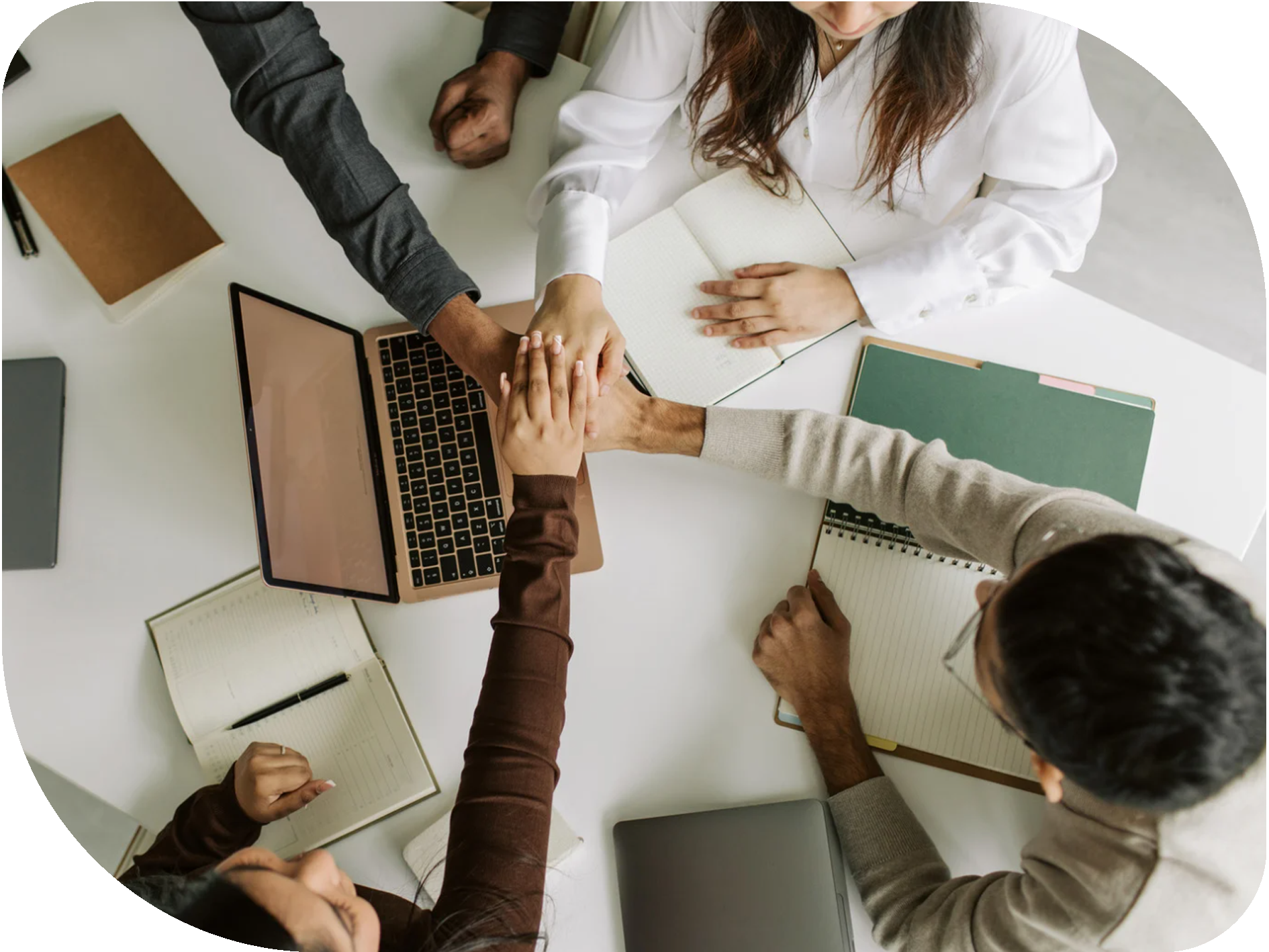 Four people at a table joining their hands together in a group hug, surrounded by notebooks, laptops, and pens.