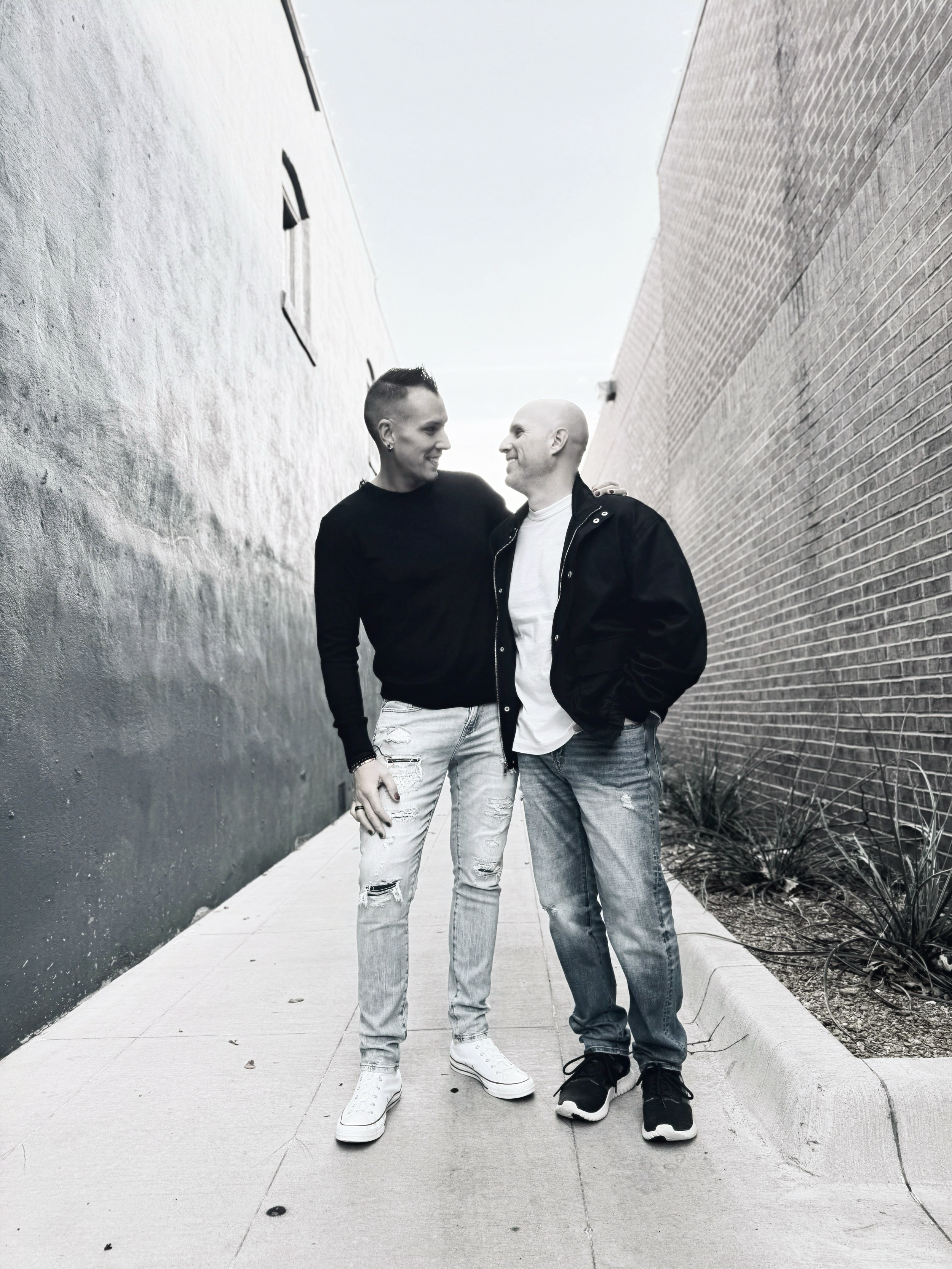 Two men standing closely in an alleyway, smiling and looking at each other, with a painted wall on one side and a brick wall on the other.