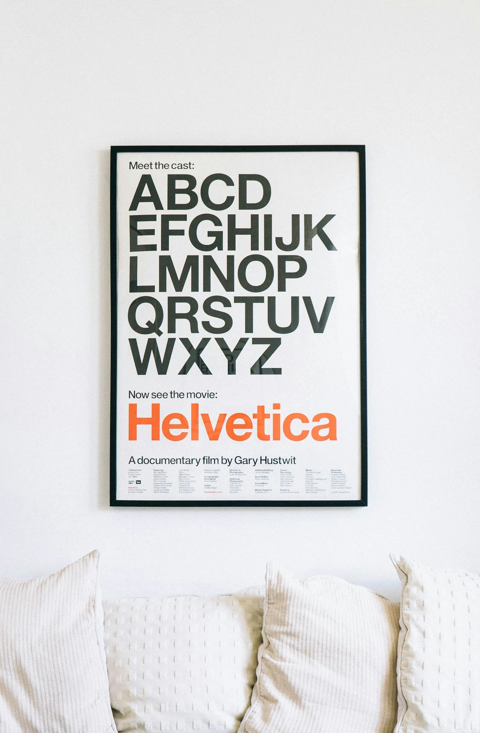 Poster on a white wall promoting the documentary film 'Helvetica' with alphabet letters and details about the film at the bottom, placed above a white textured sofa.