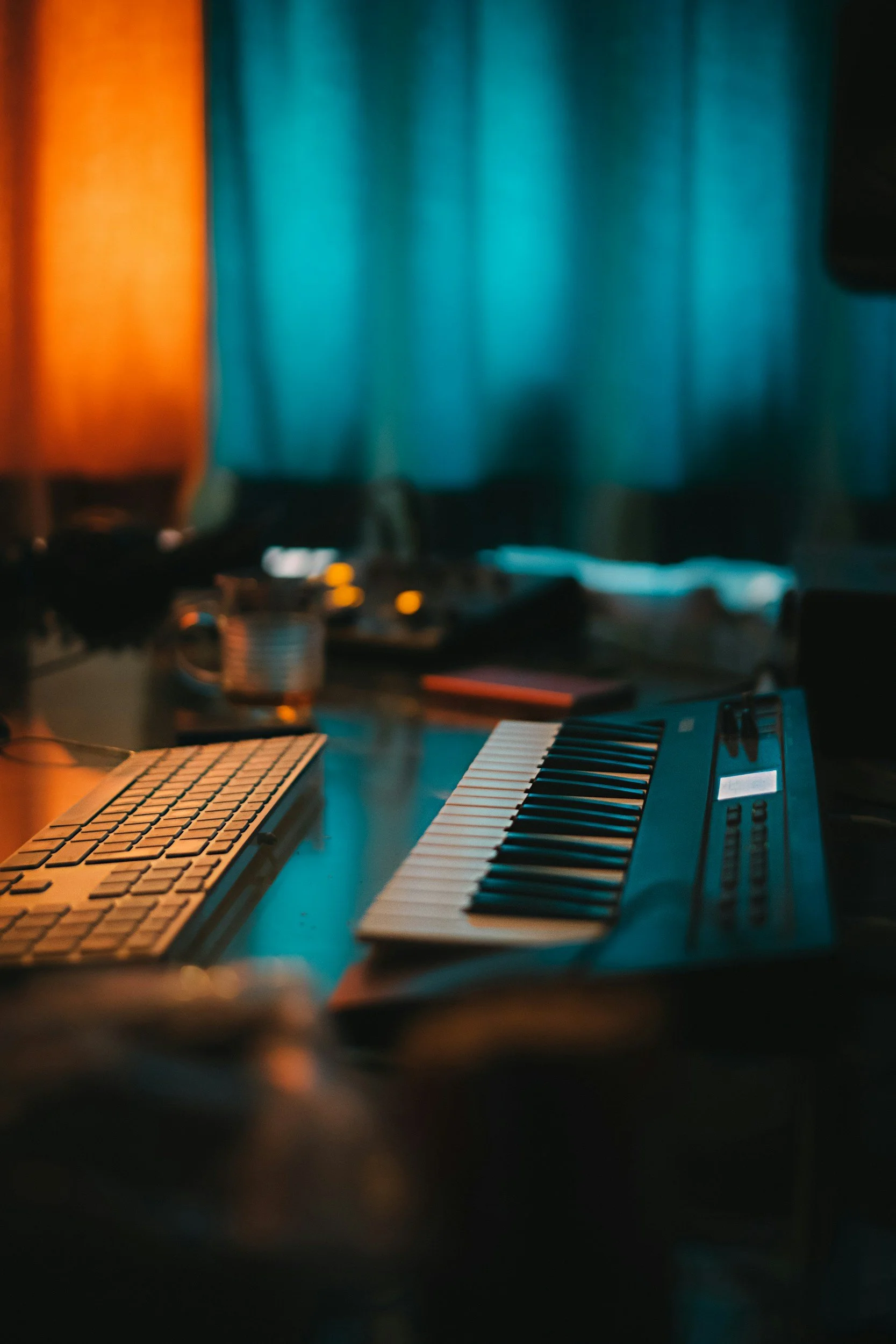 A music workspace with a MIDI keyboard and a keyboard on a dark wooden desk, dimly lit with blue and orange lighting, and a computer monitor in the background.