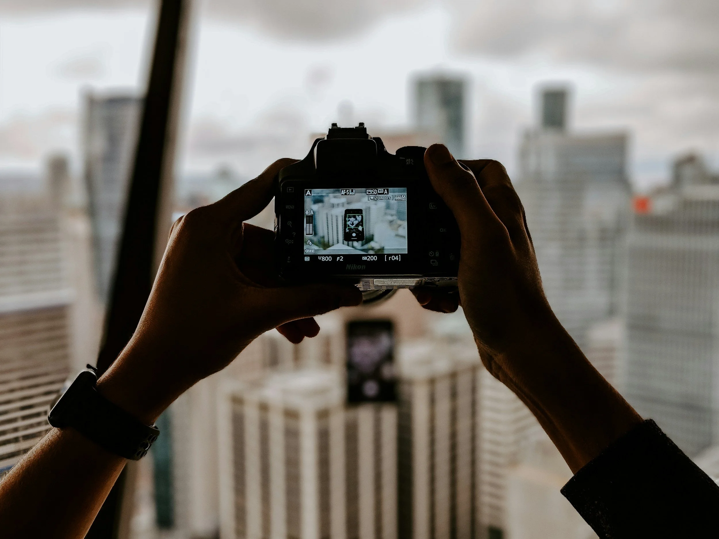 Person taking a photo of city buildings through a camera with an urban skyline in the background.