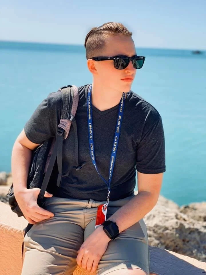 A young man with short hair, sunglasses, wearing a dark T-shirt, khaki shorts, a backpack, a lanyard around his neck, and a smartwatch, sitting on a rocky coast near the water during daytime.
