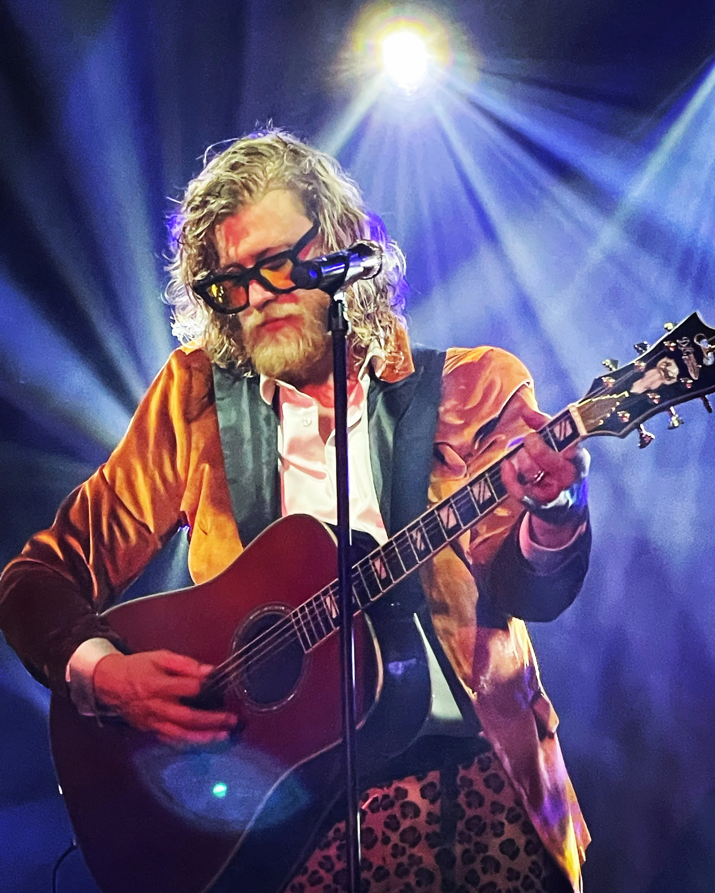 A man with wavy hair, glasses, and a beard playing an acoustic guitar on stage under bright lights.