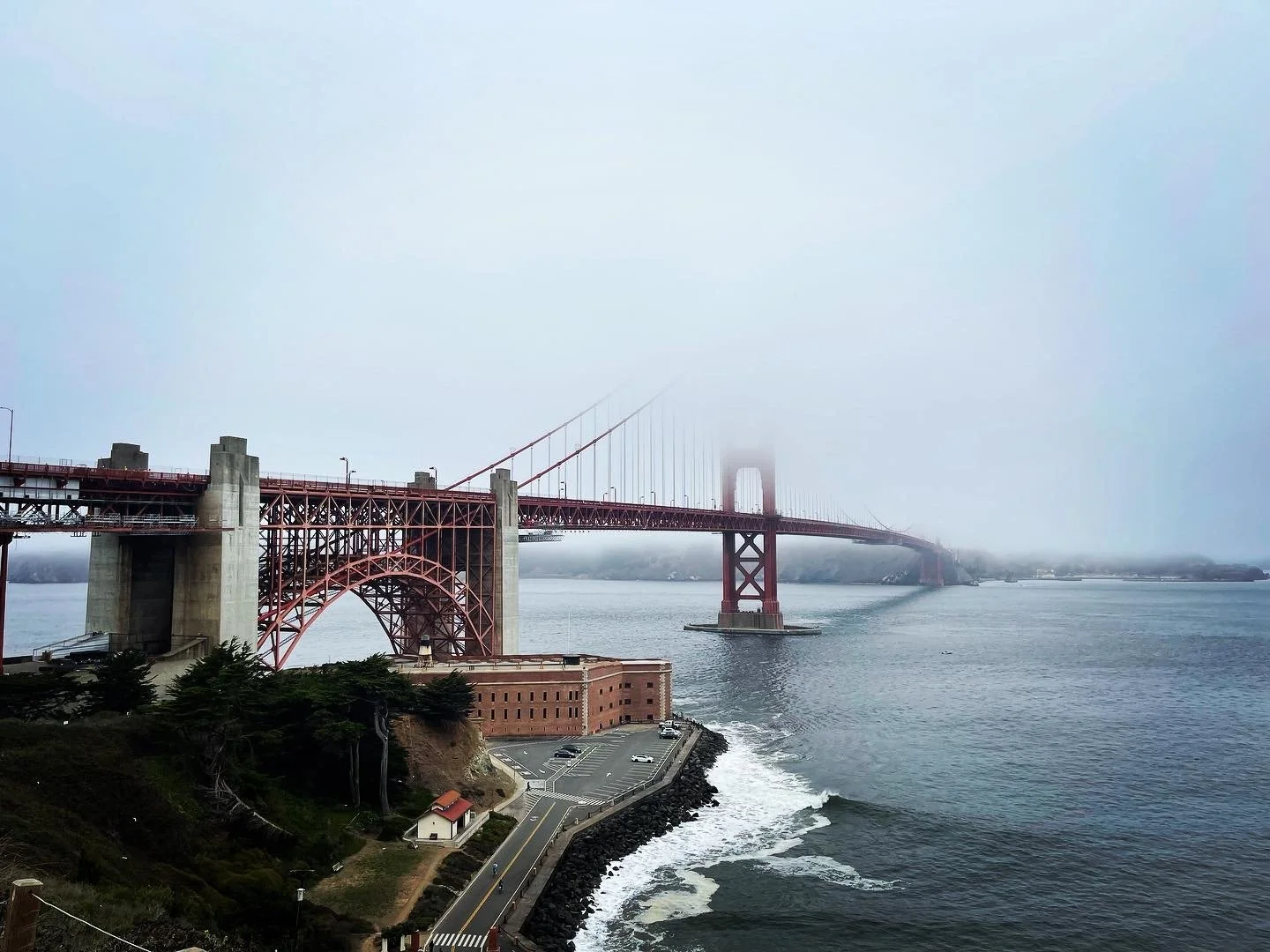 The Golden Gate Bridge extends into foggy weather over the water, with a small parking lot and trees in the foreground.