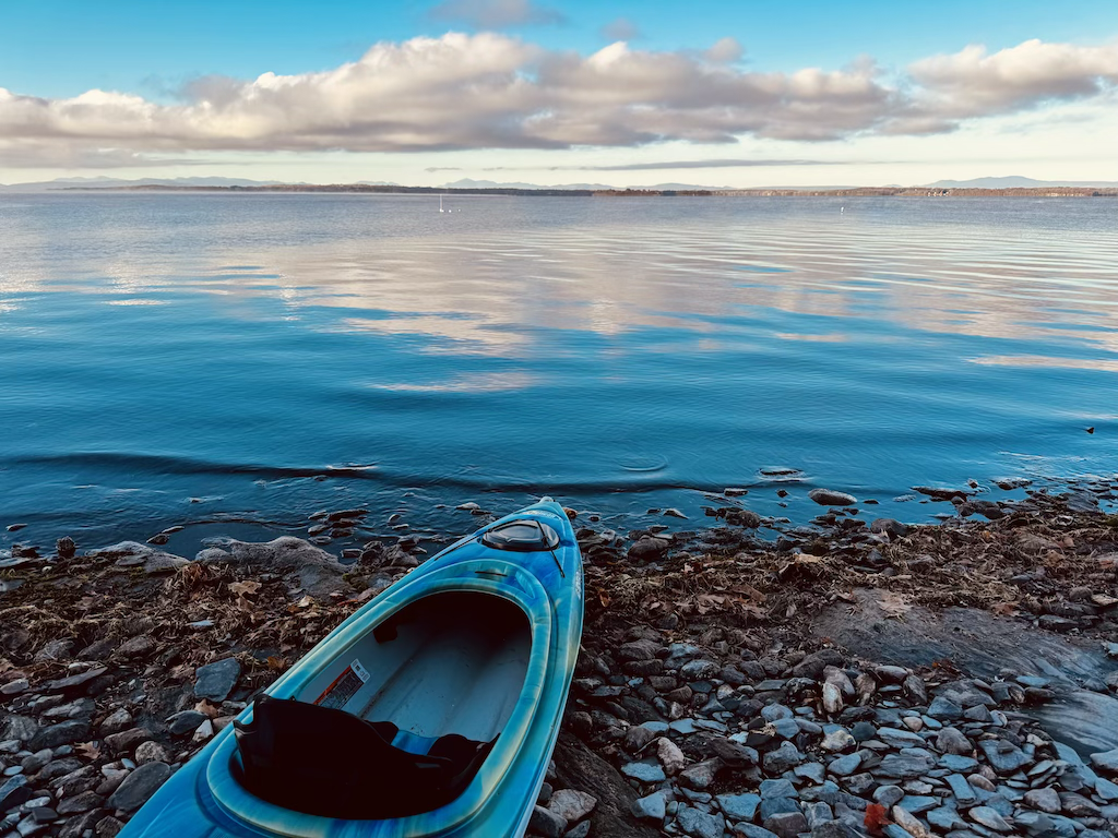 A blue kayak resting on a rocky shoreline by a calm body of water with a distant landmass and a partly cloudy sky.