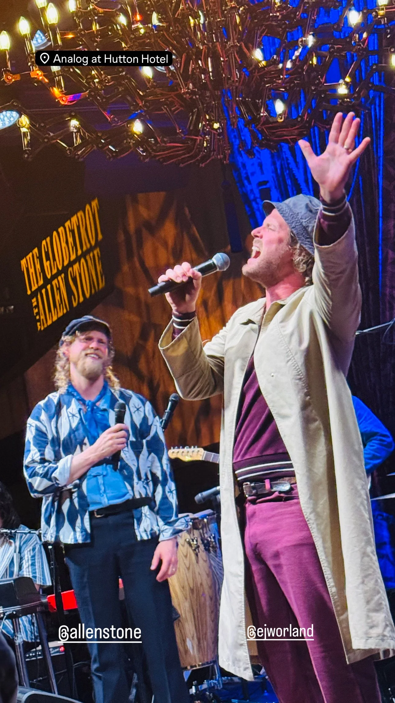 Two performers singing on stage at the Analog at Hutton Hotel. One man with curly hair, a beard, wearing a patterned shirt and black hat, holds a microphone. The other man with curly hair, wearing a bucket hat, beige trench coat, maroon pants, and a 