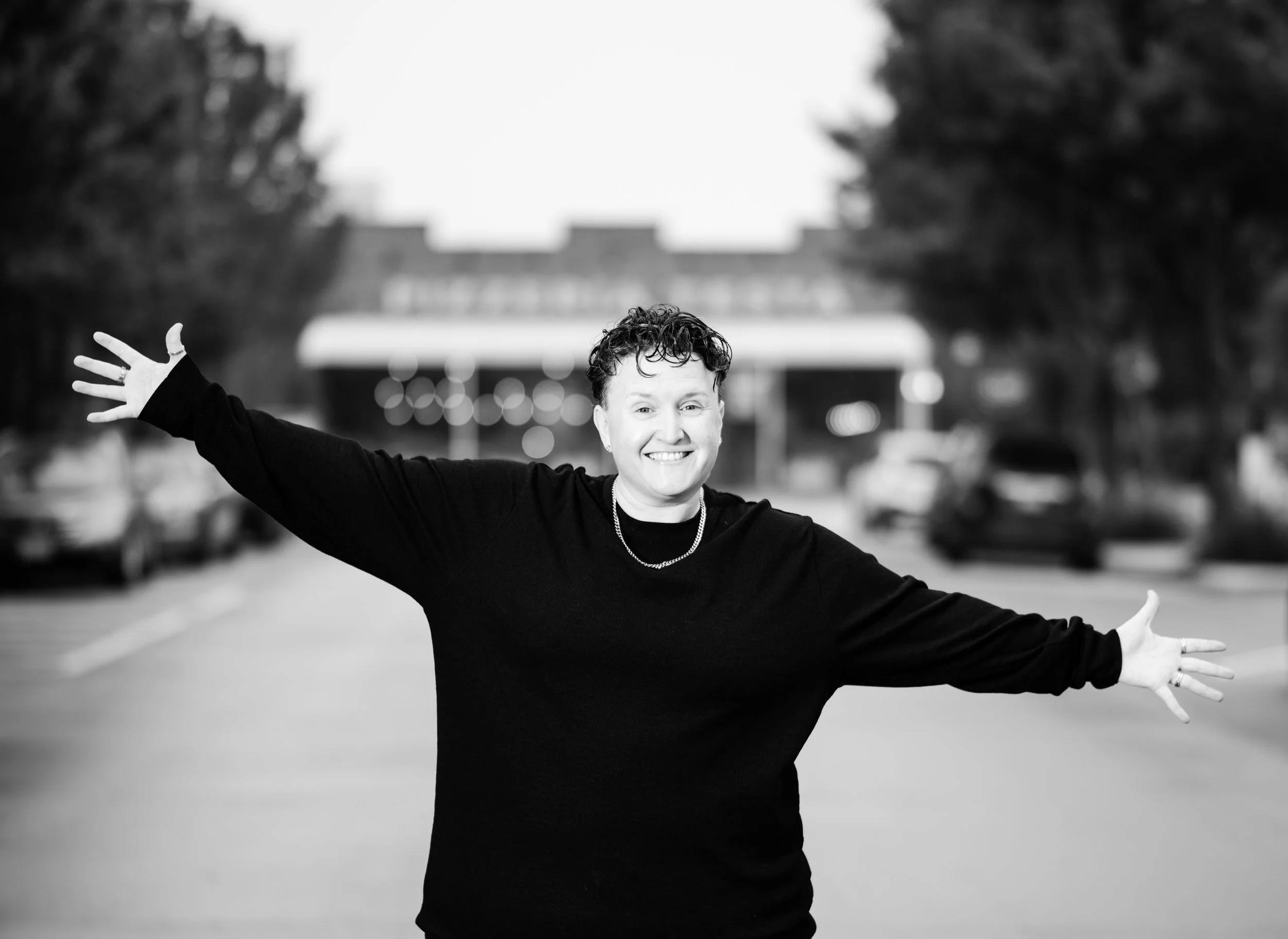 A smiling person with short curly hair standing in the middle of a street with arms outstretched, wearing a black long-sleeve shirt, with parked cars and trees in the background.