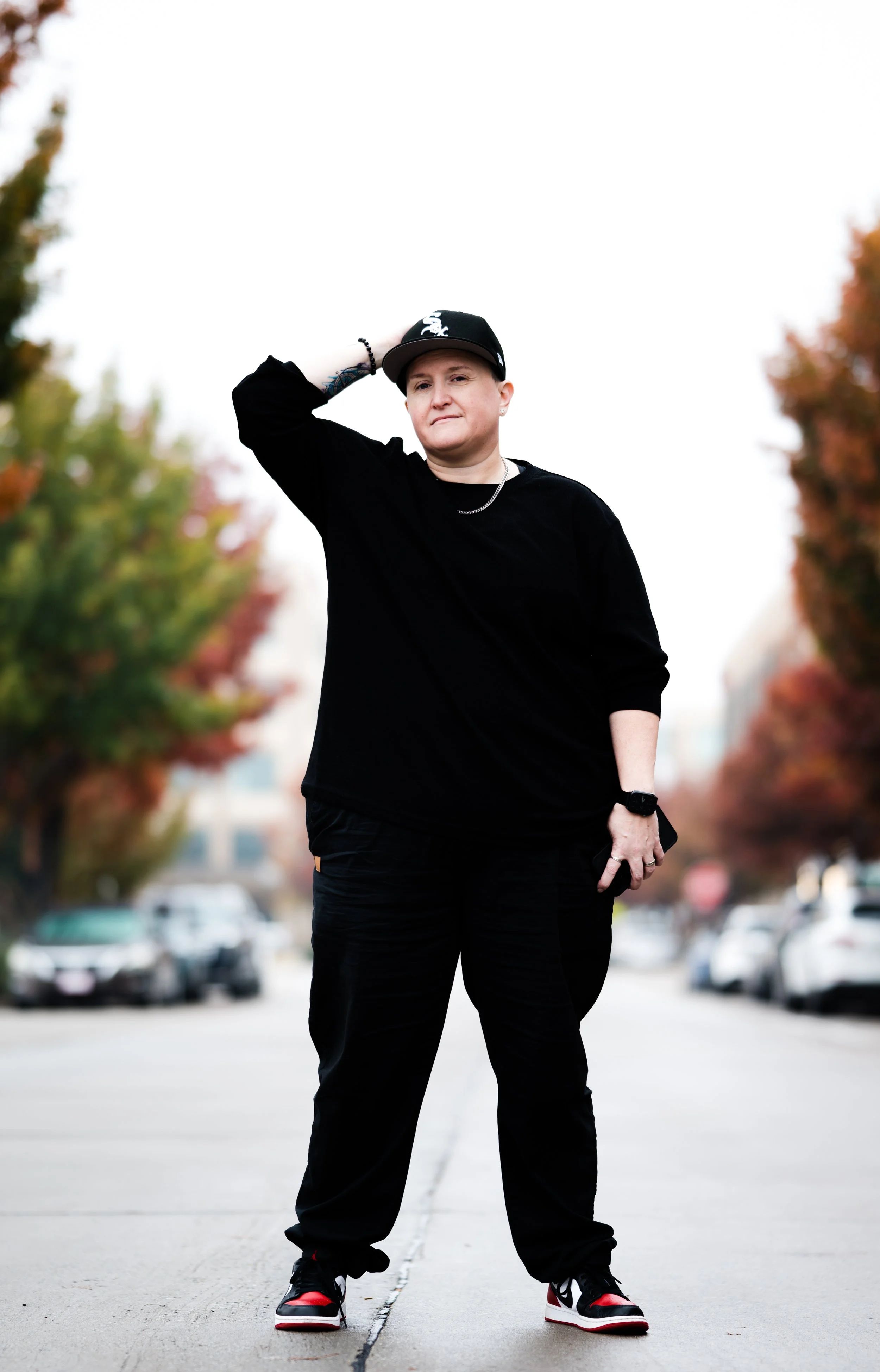 A person standing in the middle of a city street during fall, wearing black clothing, a baseball cap, and sneakers, with trees showing fall colors in the background.