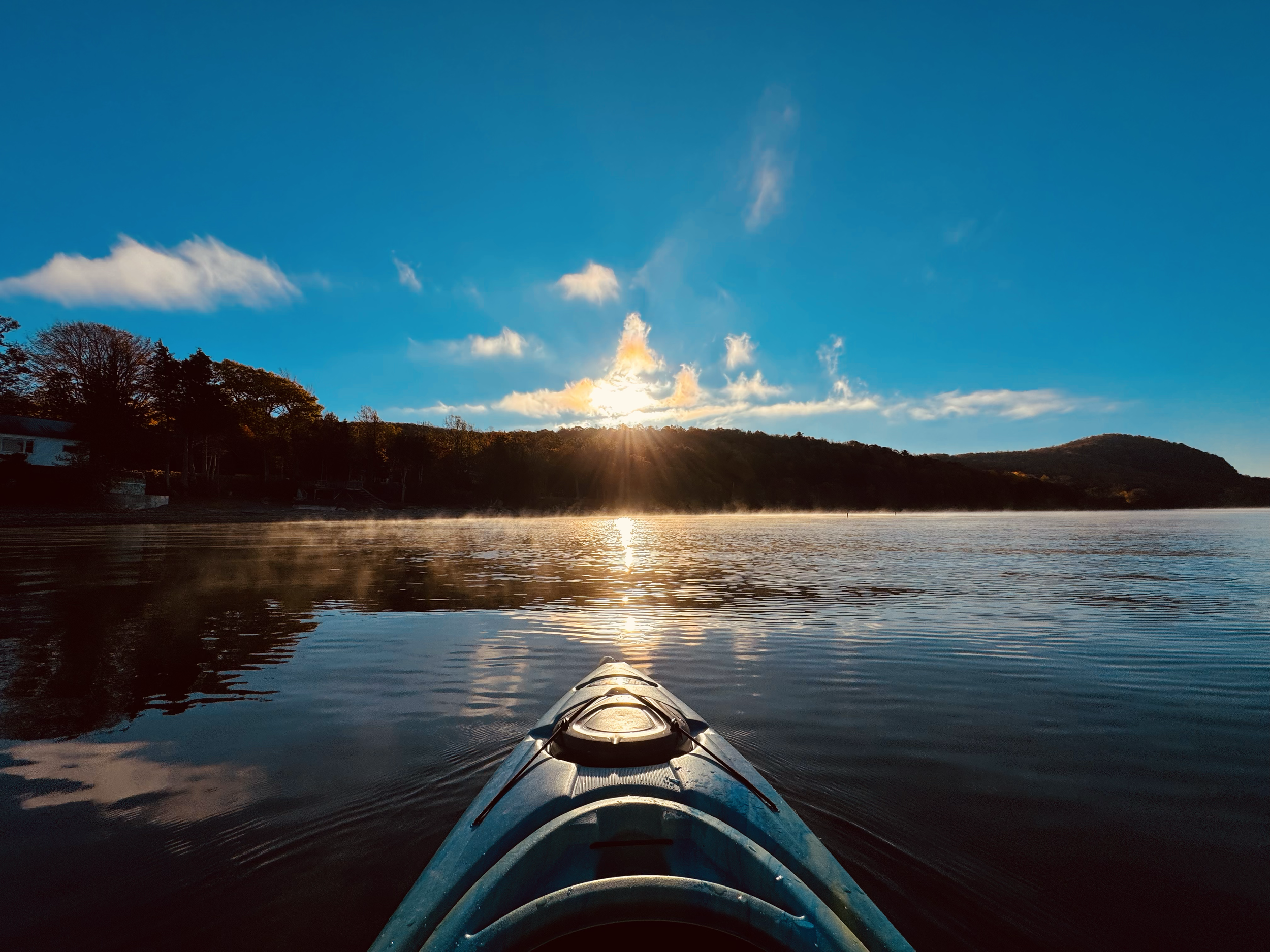 View from a kayak on a calm lake during sunrise, with a clear sky, some clouds, and a forested shoreline in the distance.