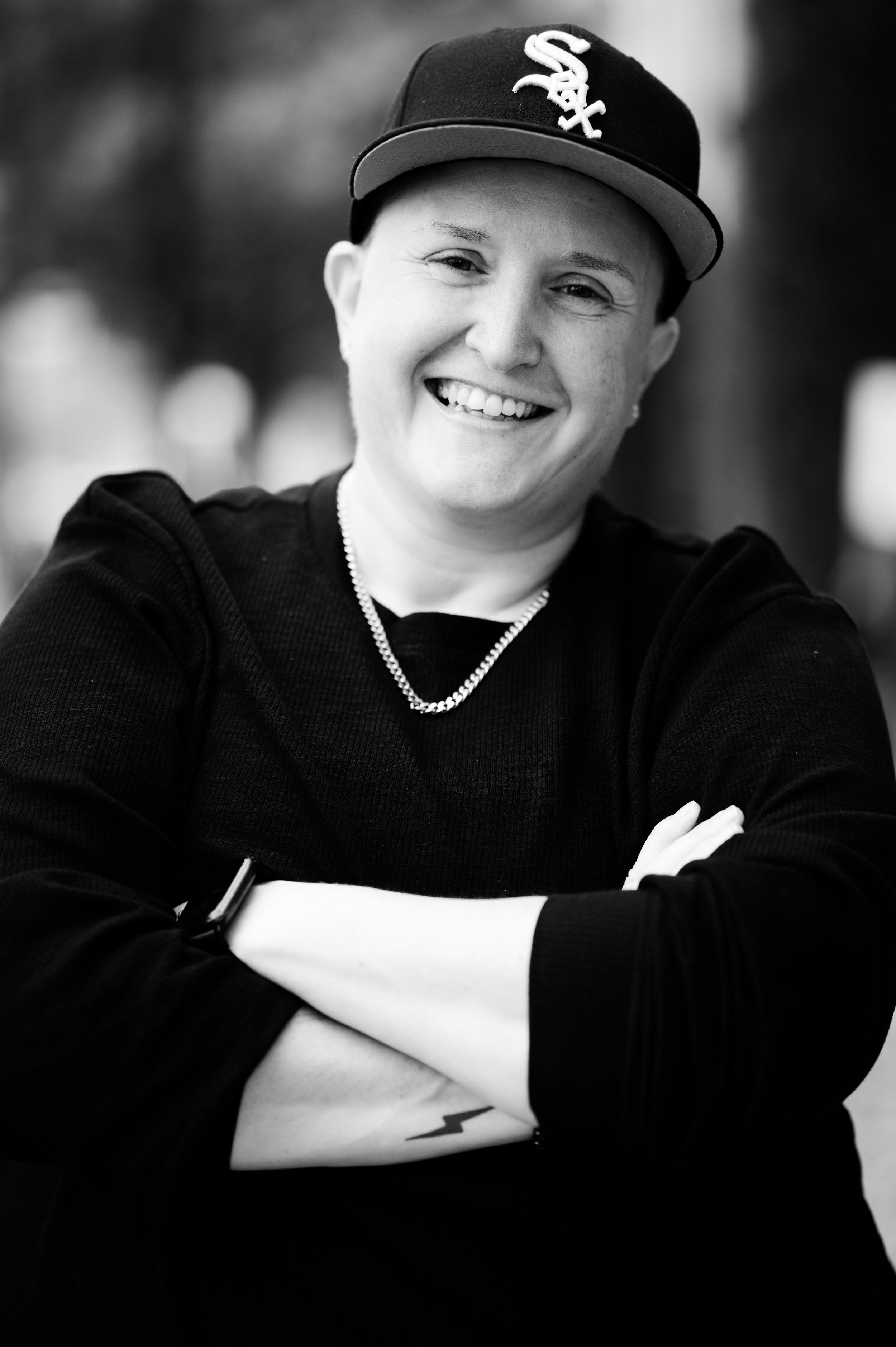 Black and white photo of a smiling person wearing a baseball cap with a Chicago White Sox logo, black long-sleeve shirt, chain necklace, and with arms crossed.