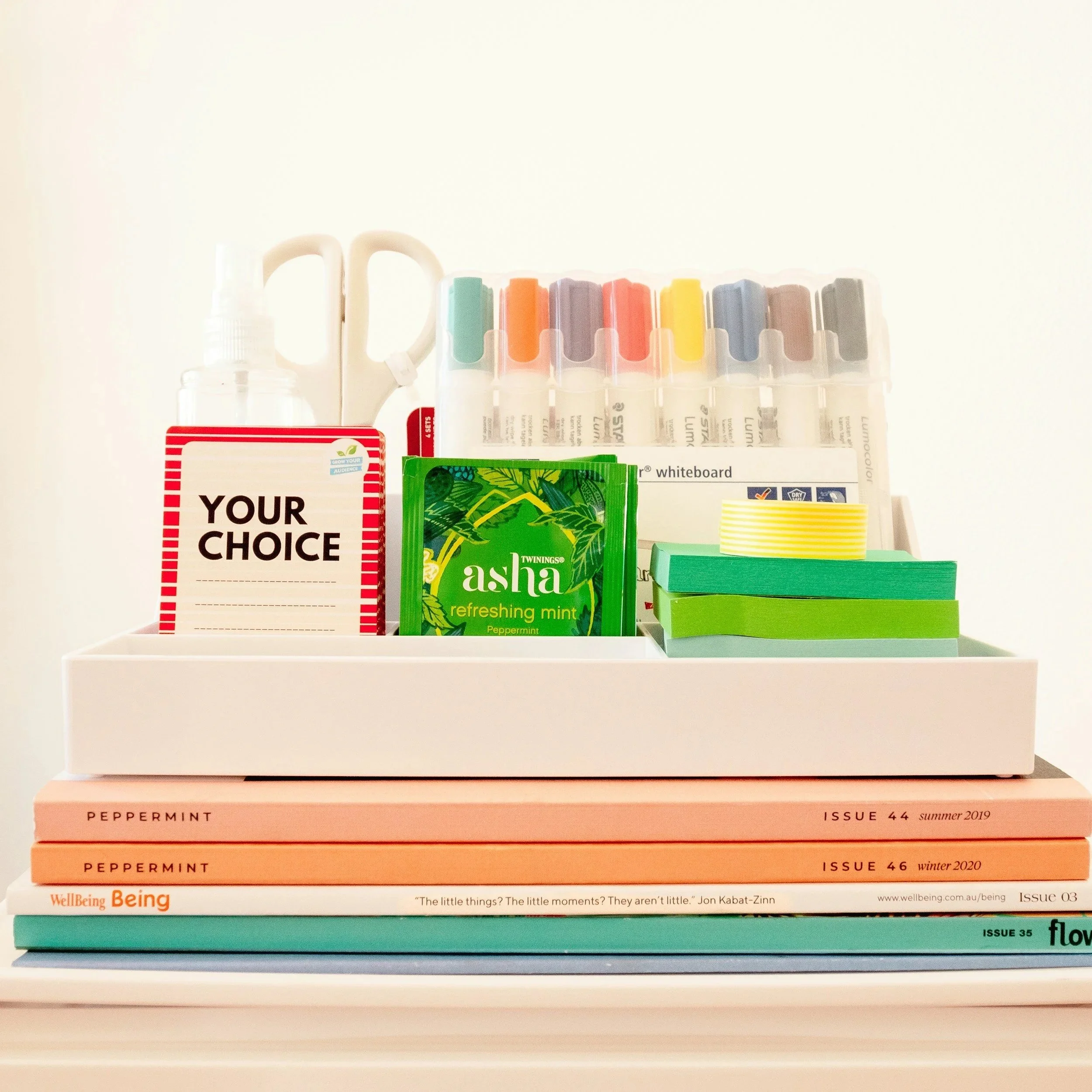 A desk organizer with office supplies, including books labeled 'Peppermint' and 'WellBeing Being,' a pack of Asha mints, sticky notes, whiteboard markers, a pair of scissors, a spray bottle, and a clipboard, arranged on a white surface with a plain background.