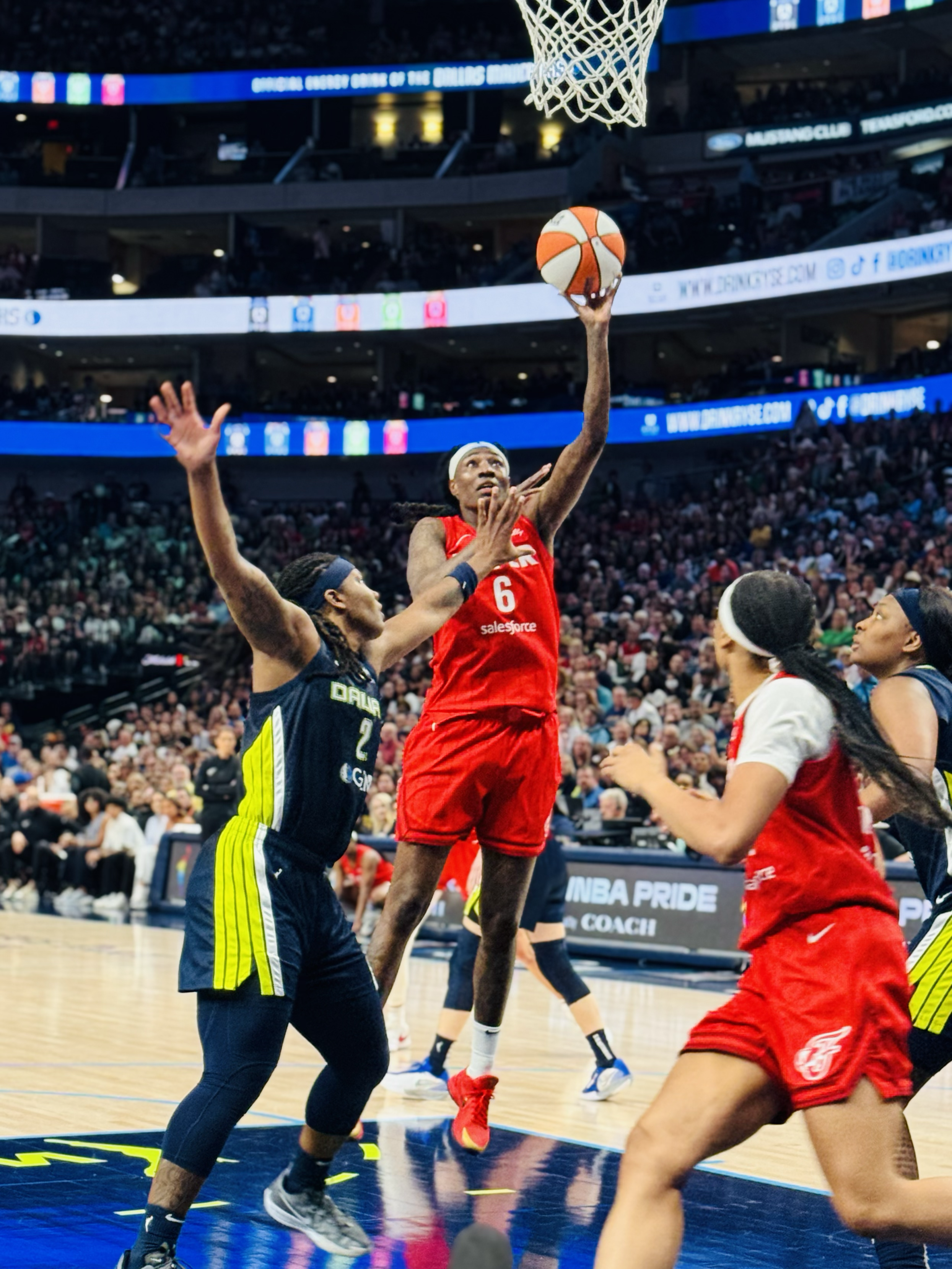A basketball game with players from different teams competing near the basket. A player in a red jersey, number 6, is jumping to make a shot, while a player in a black jersey, number 2, attempts to block. Other players and an audience are visible in 