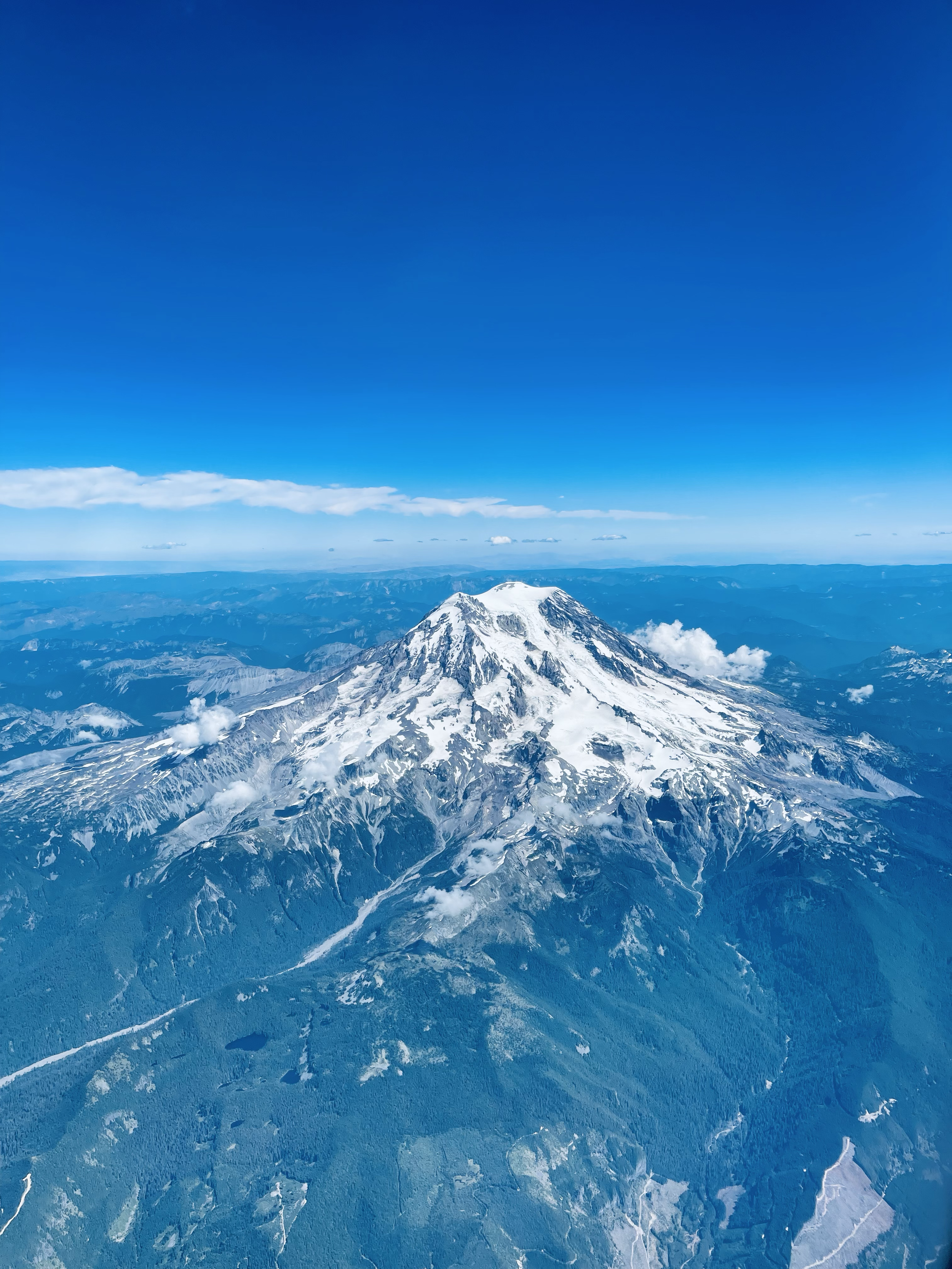 A snow-capped mountain under a blue sky with some clouds and distant mountain ranges.
